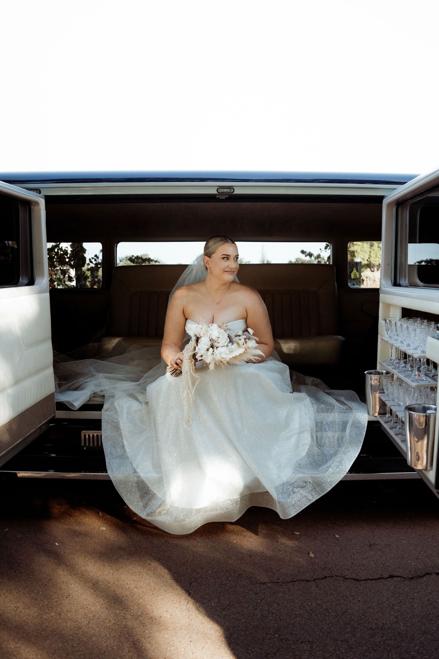 Bride in white strapless gown holding bouquet seated in back of limousine on wedding day.