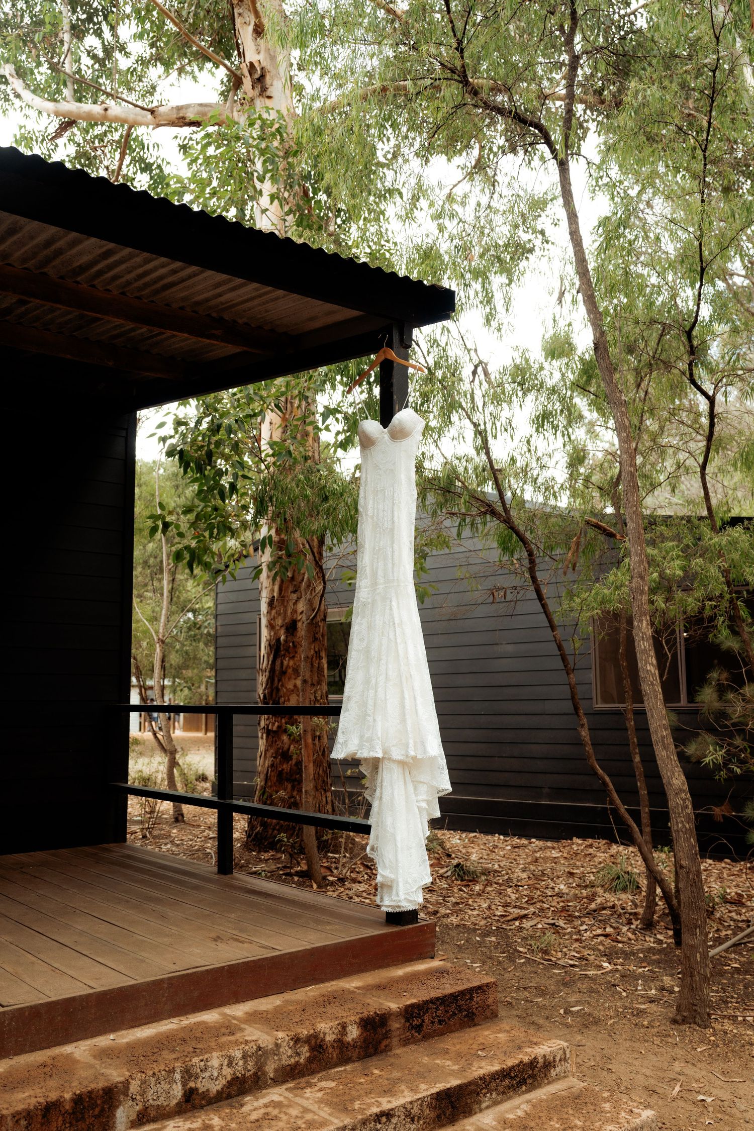 White wedding dress hanging outdoors on a rustic wooden deck surrounded by eucalyptus trees.