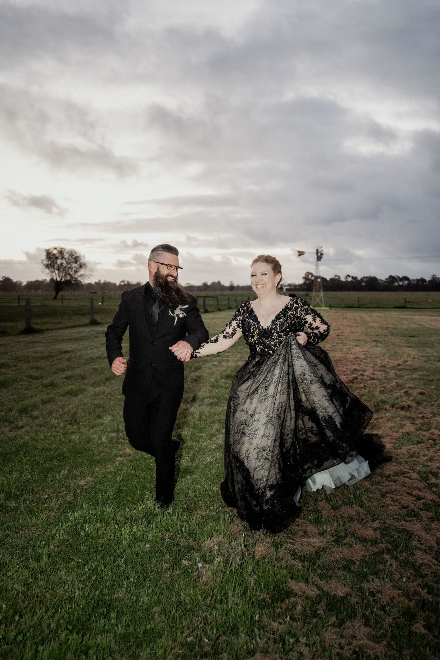 Couple in black wedding attire running through green field at dusk, bride in black lace gown, groom in black suit.