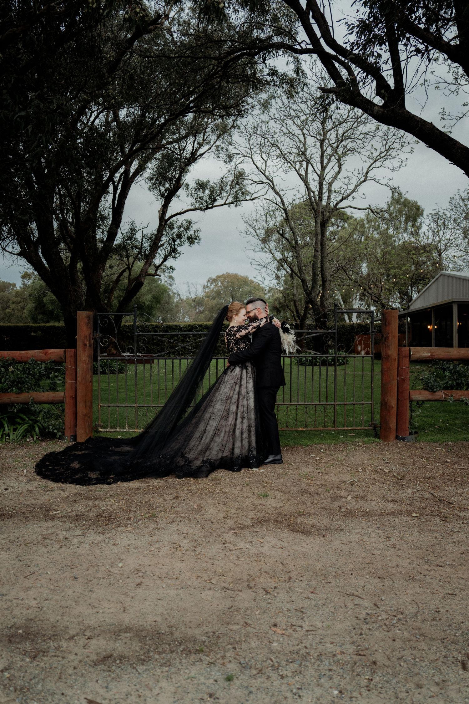 Couple kissing in black wedding attire near rustic farm gate surrounded by lush green trees on overcast day.