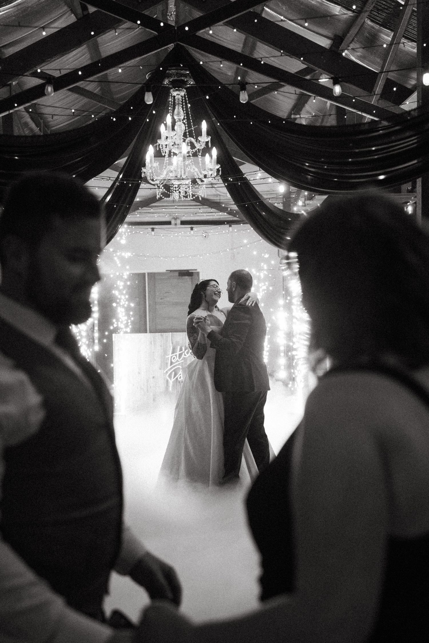 Bride and groom share first dance surrounded by fog and fairy lights under a chandelier in black and white.