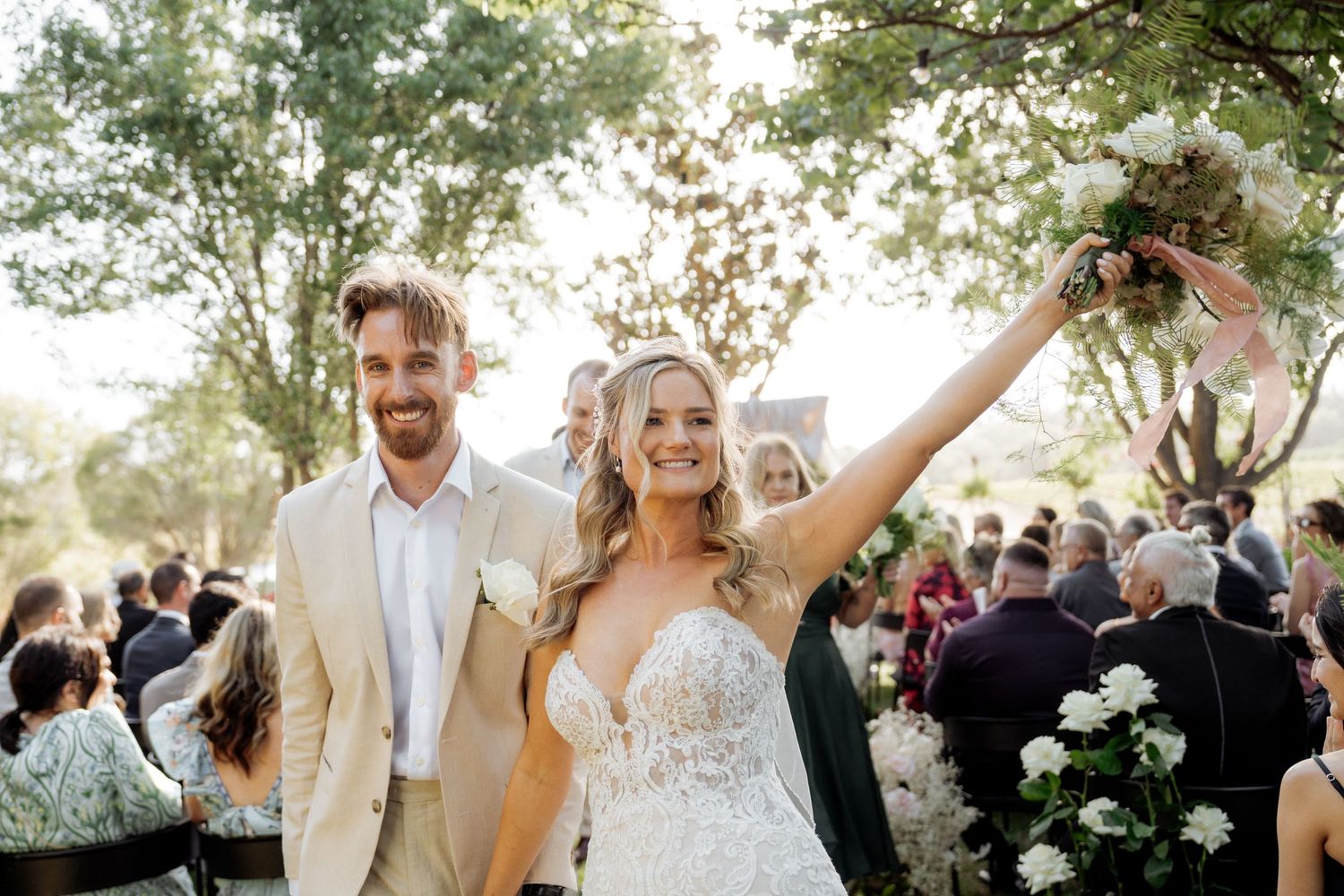 Joyful newlyweds walk down the aisle, bride in lace gown raising bouquet, groom in tan suit, outdoor wedding ceremony.