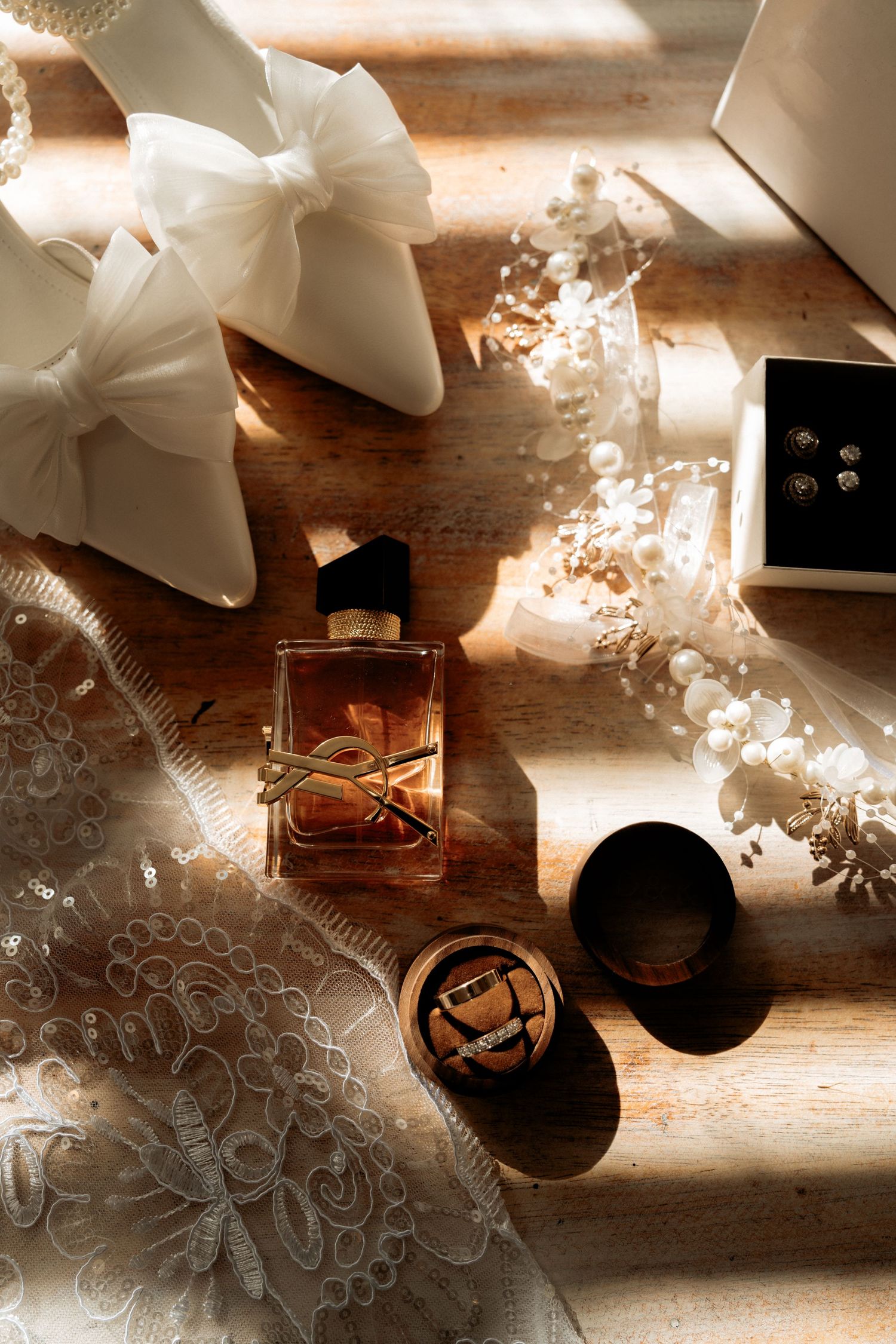 Bridal flat lay with YSL perfume, white heels, pearl headpiece, earrings, and lace on rustic wood in warm sunlight.