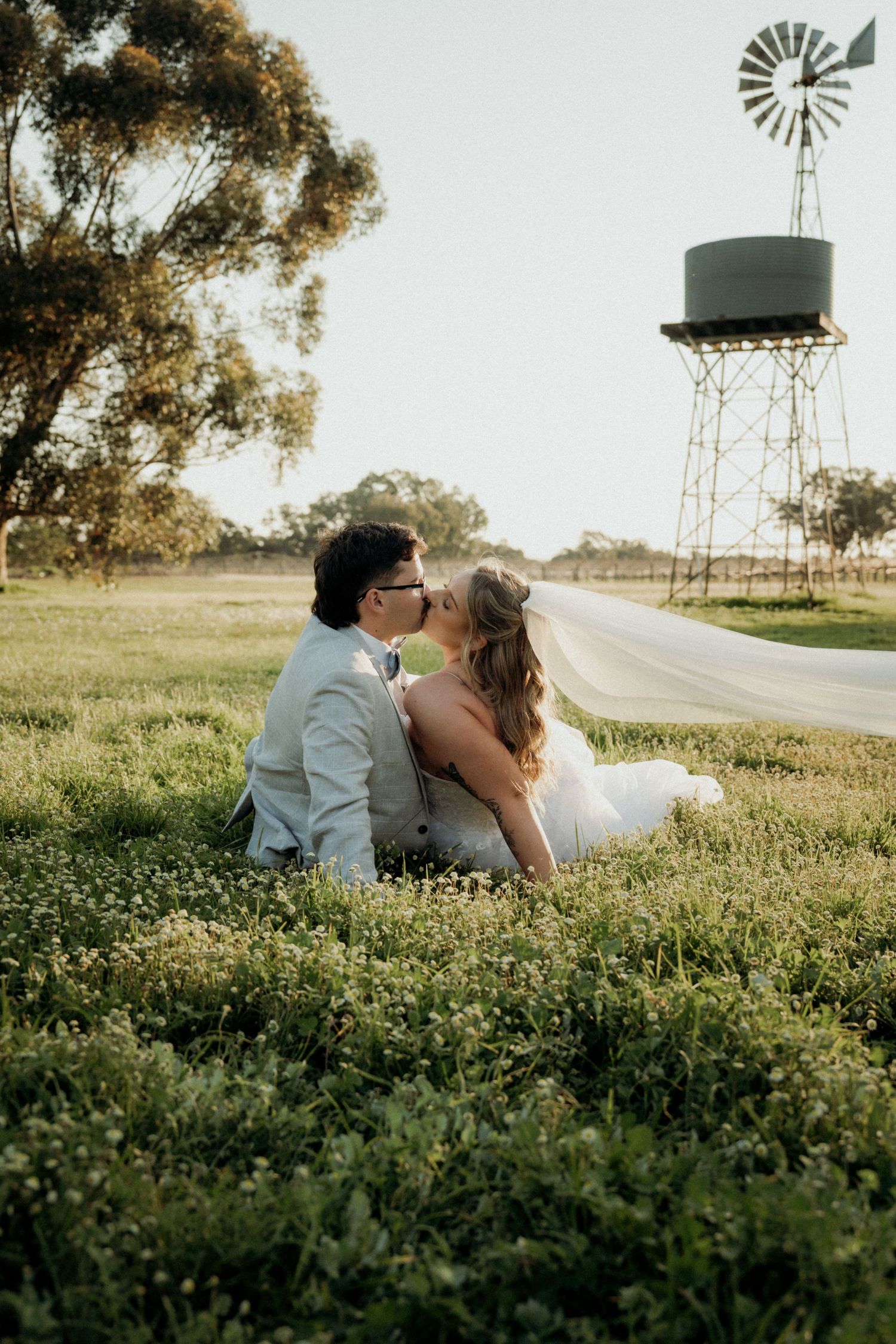 Newlywed couple kissing on grass at golden hour, with a rustic windmill and water tower in the background.