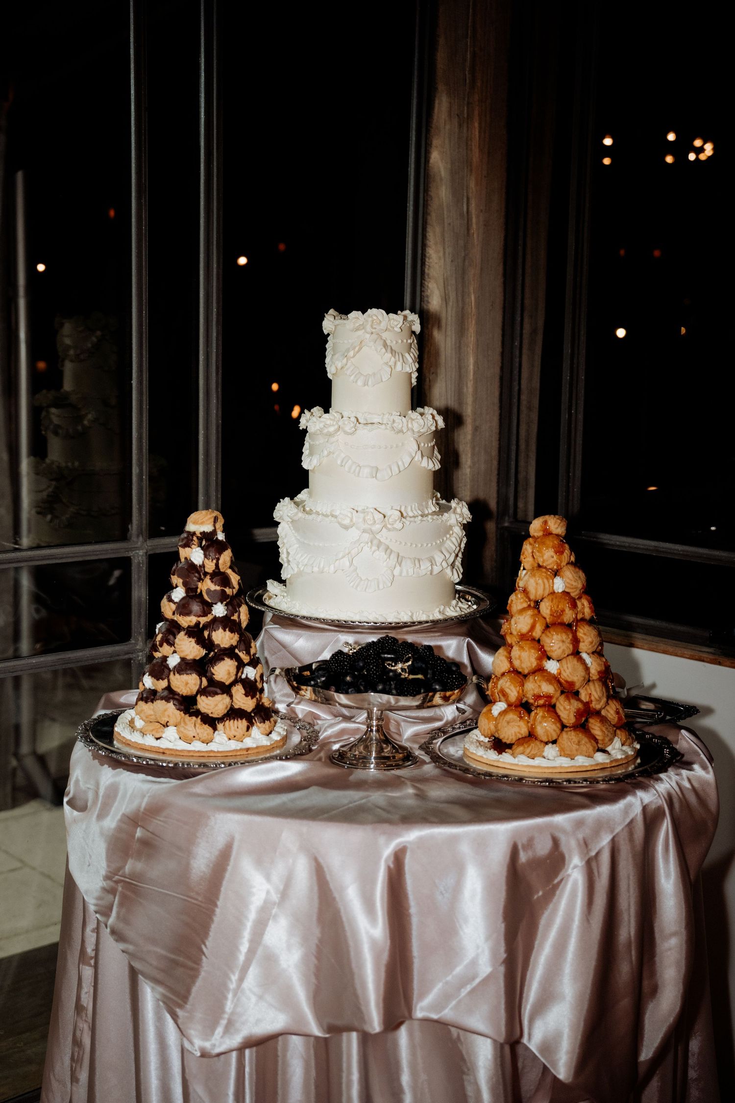 Elegant three-tier white wedding cake displayed with two croquembouche towers on a satin-draped table at a night reception.