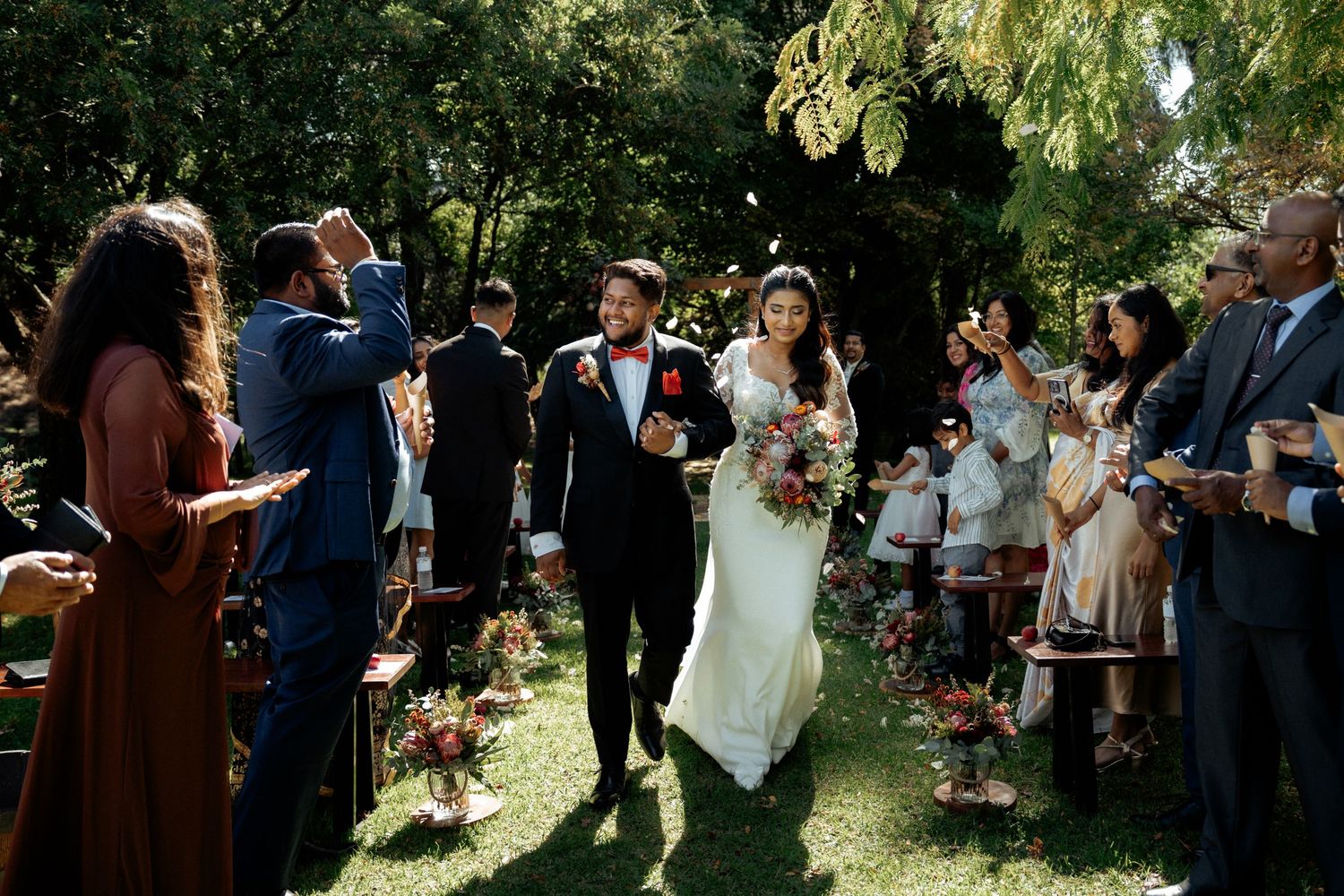 Bride and groom walk down outdoor aisle as guests celebrate with confetti at a garden wedding ceremony.