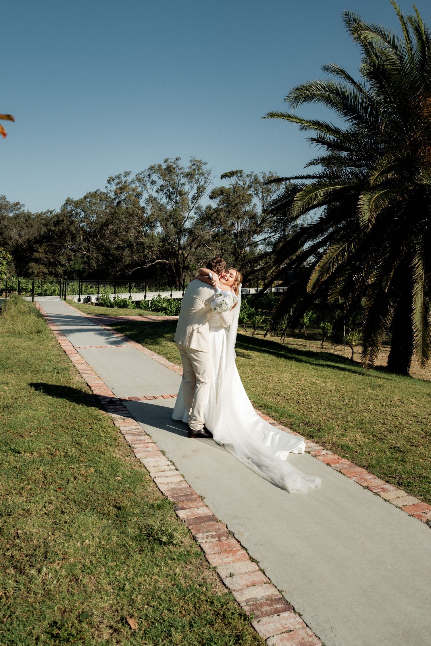Bride and groom sharing a romantic kiss on a garden pathway lined with palm trees on a sunny wedding day.