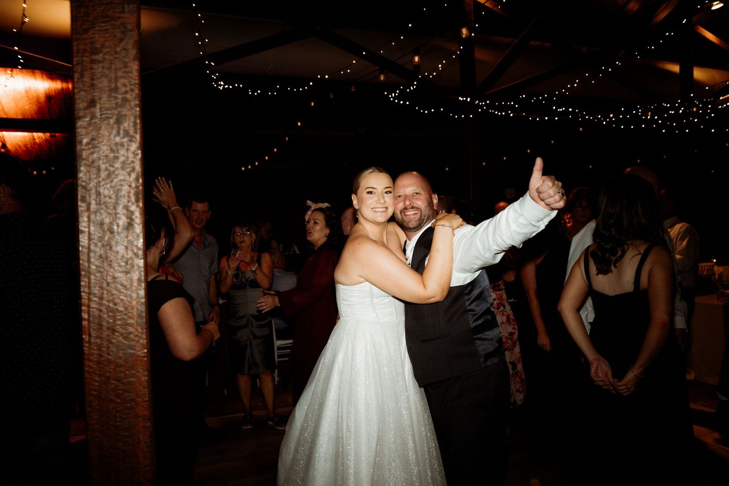 Bride and groom dancing joyfully at their wedding reception with guests and string lights in the background.