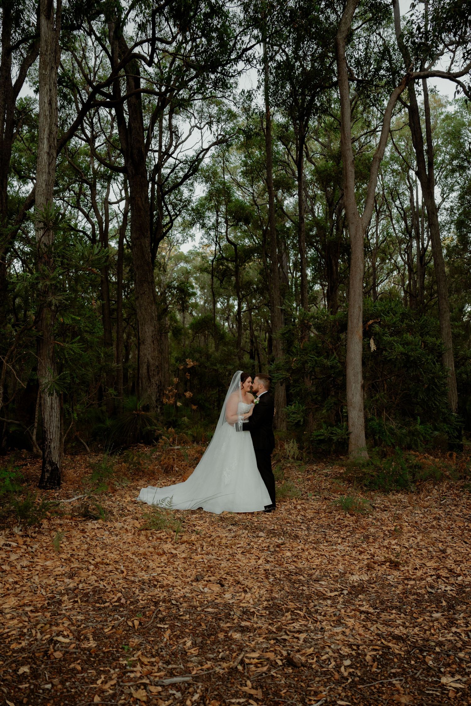 Bride and groom share a kiss in a forest clearing, surrounded by tall trees and a carpet of autumn leaves.