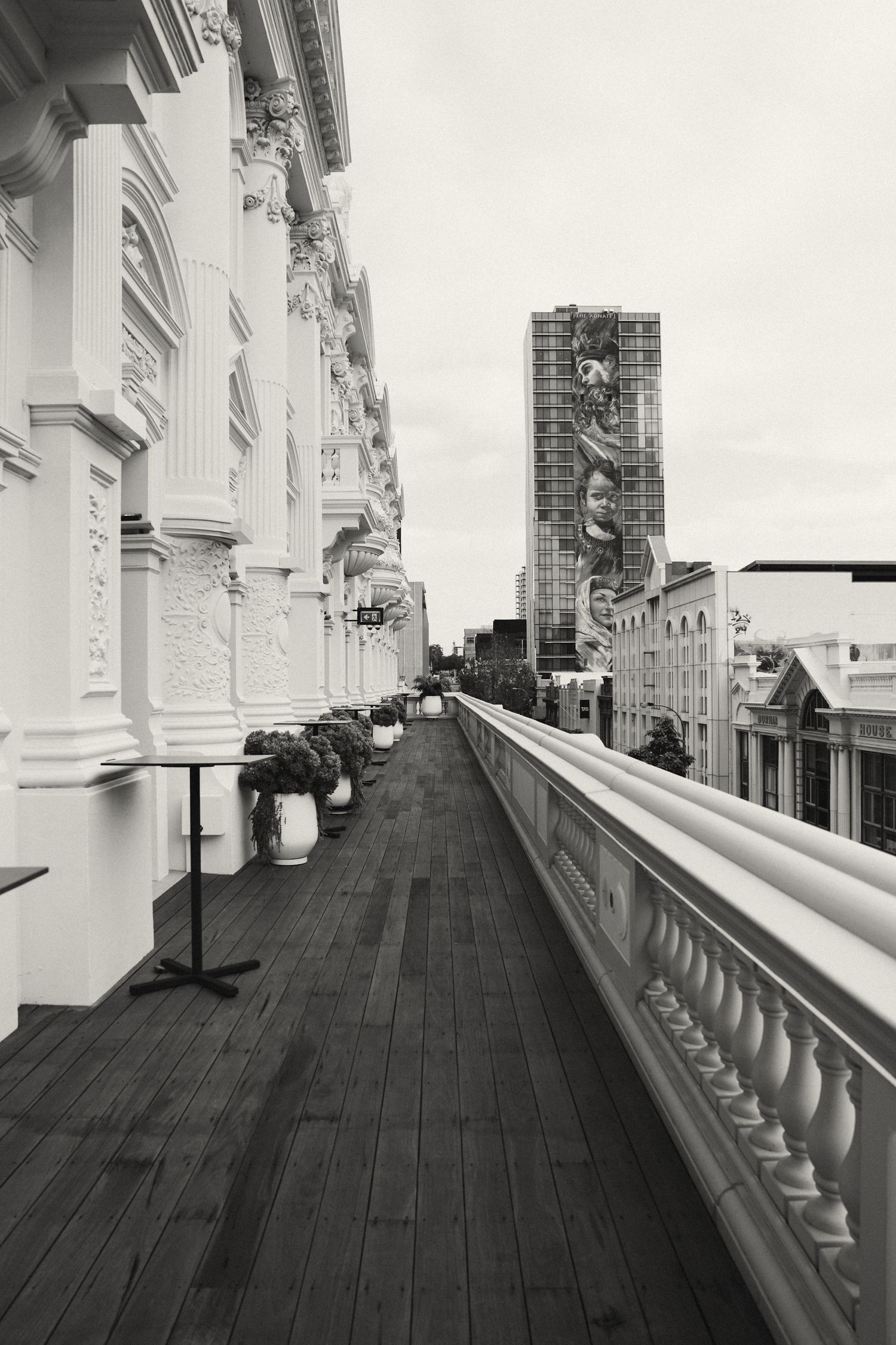A black and white view of an ornate hotel balcony with white columns and wooden deck overlooking a city street and tower.