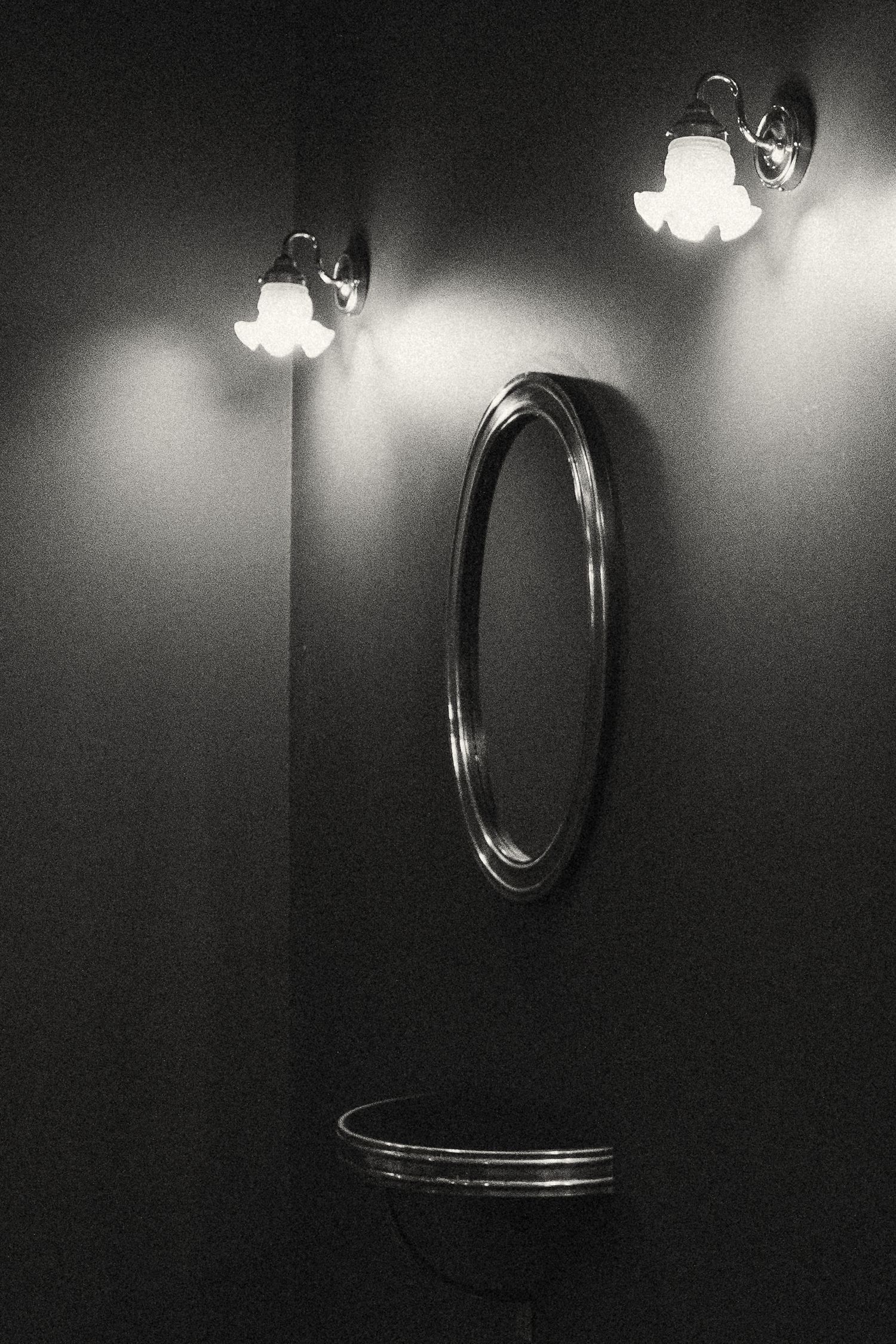 A dramatic black and white photograph series of an oval mirror mounted on a dark wall with a vintage wall sconce light above.