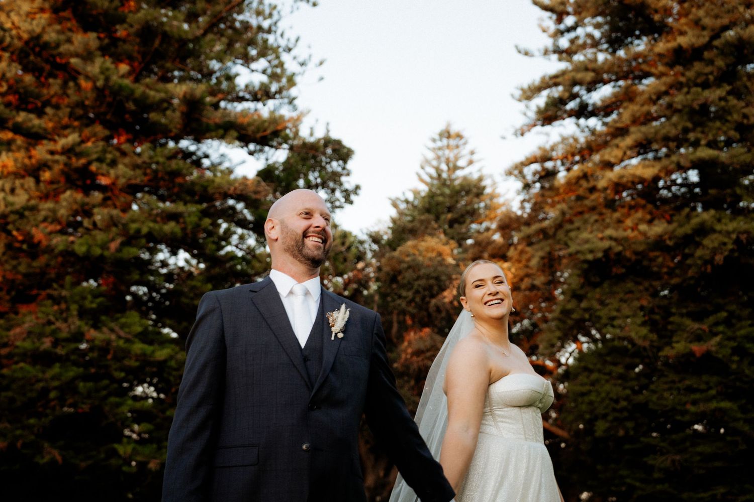 A bride and groom hold hands smiling outdoors with autumn foliage trees in background on their wedding day.