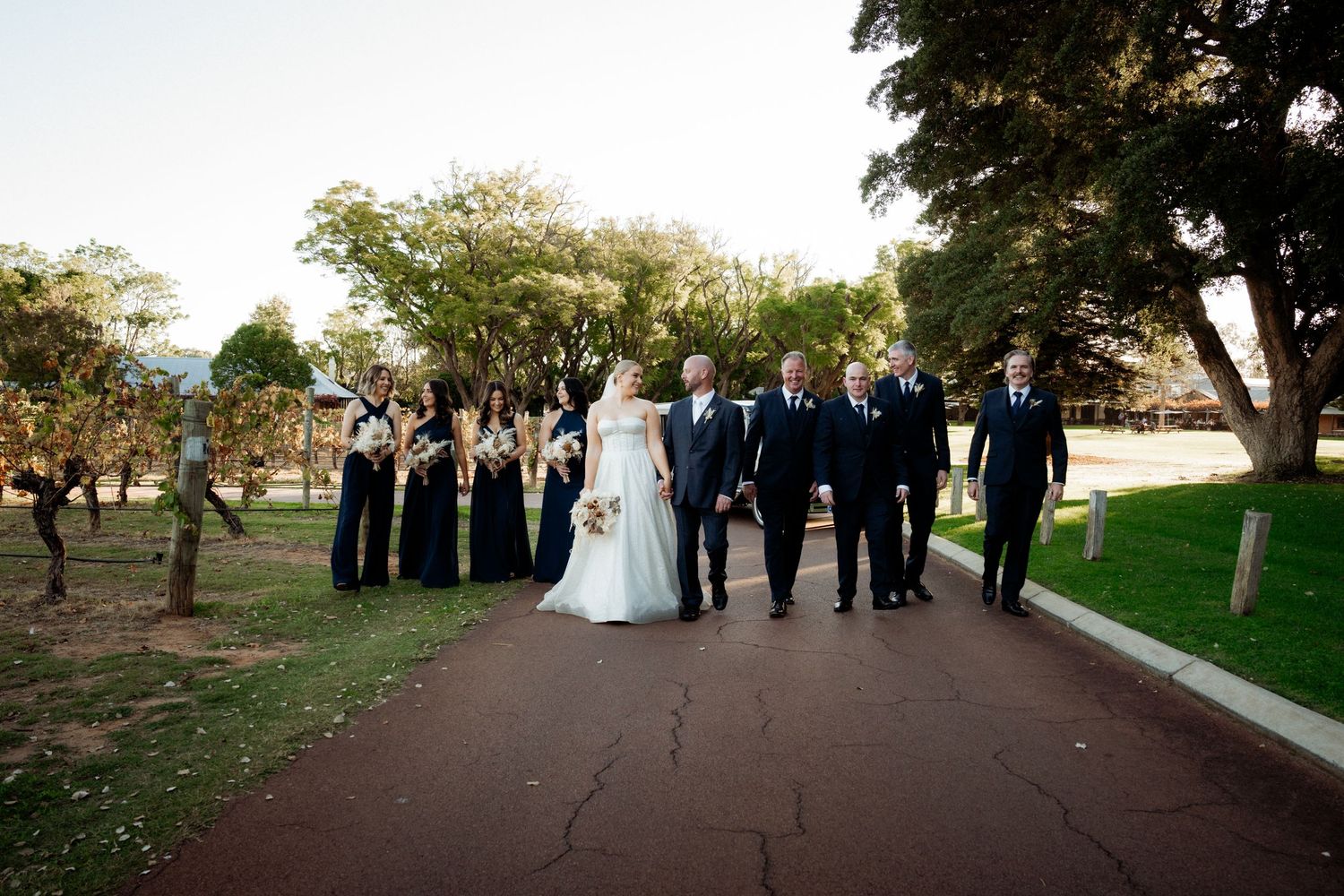 Wedding party walks along a path outdoors, bride in white gown with bridesmaids in black dresses and groomsmen in suits.