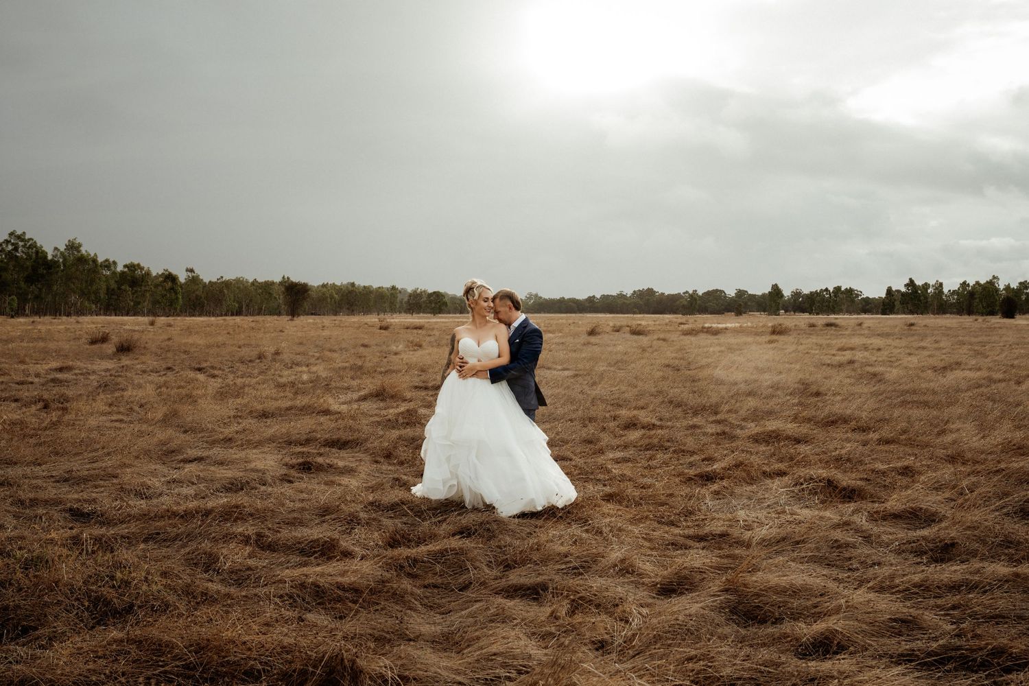 Bride and groom embrace in a golden field under dramatic cloudy skies on their wedding day.