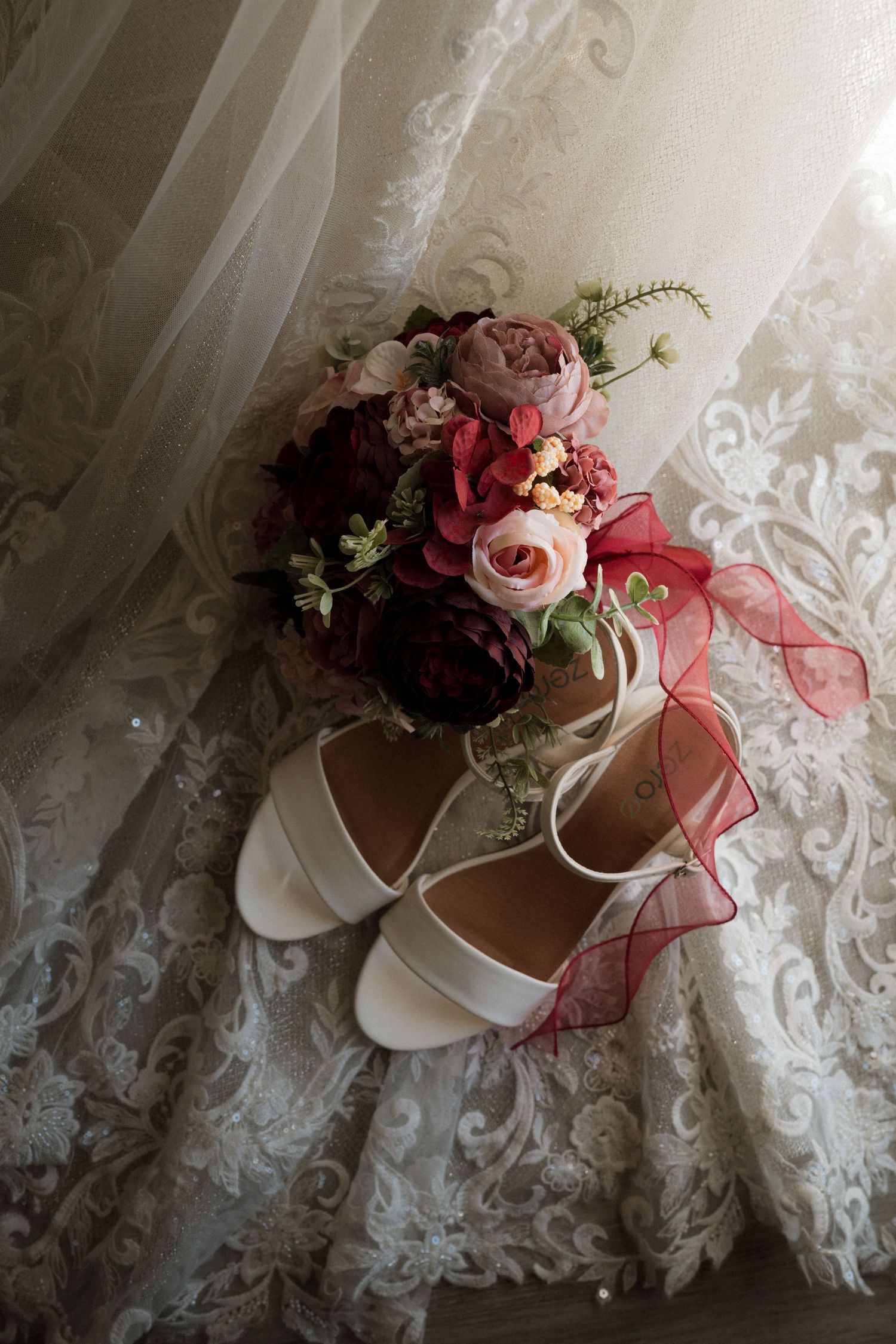 White bridal heels and a burgundy and pink rose bouquet with red ribbon rest on lace wedding dress fabric.