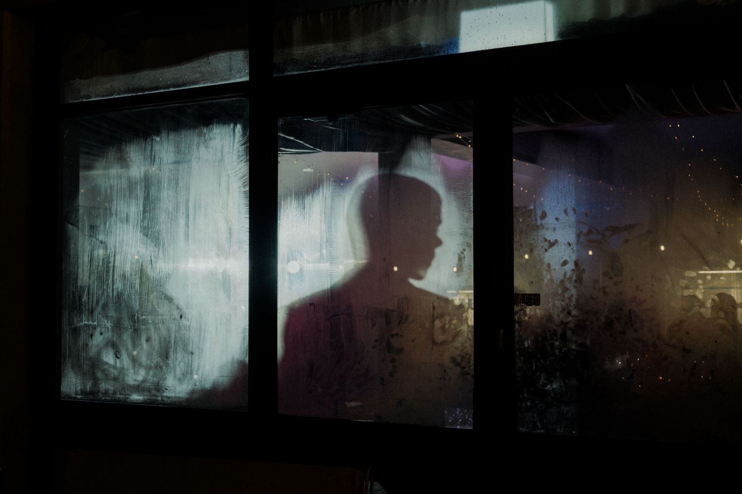 Silhouette of a person visible through a foggy, rain-streaked window at night with dim light.