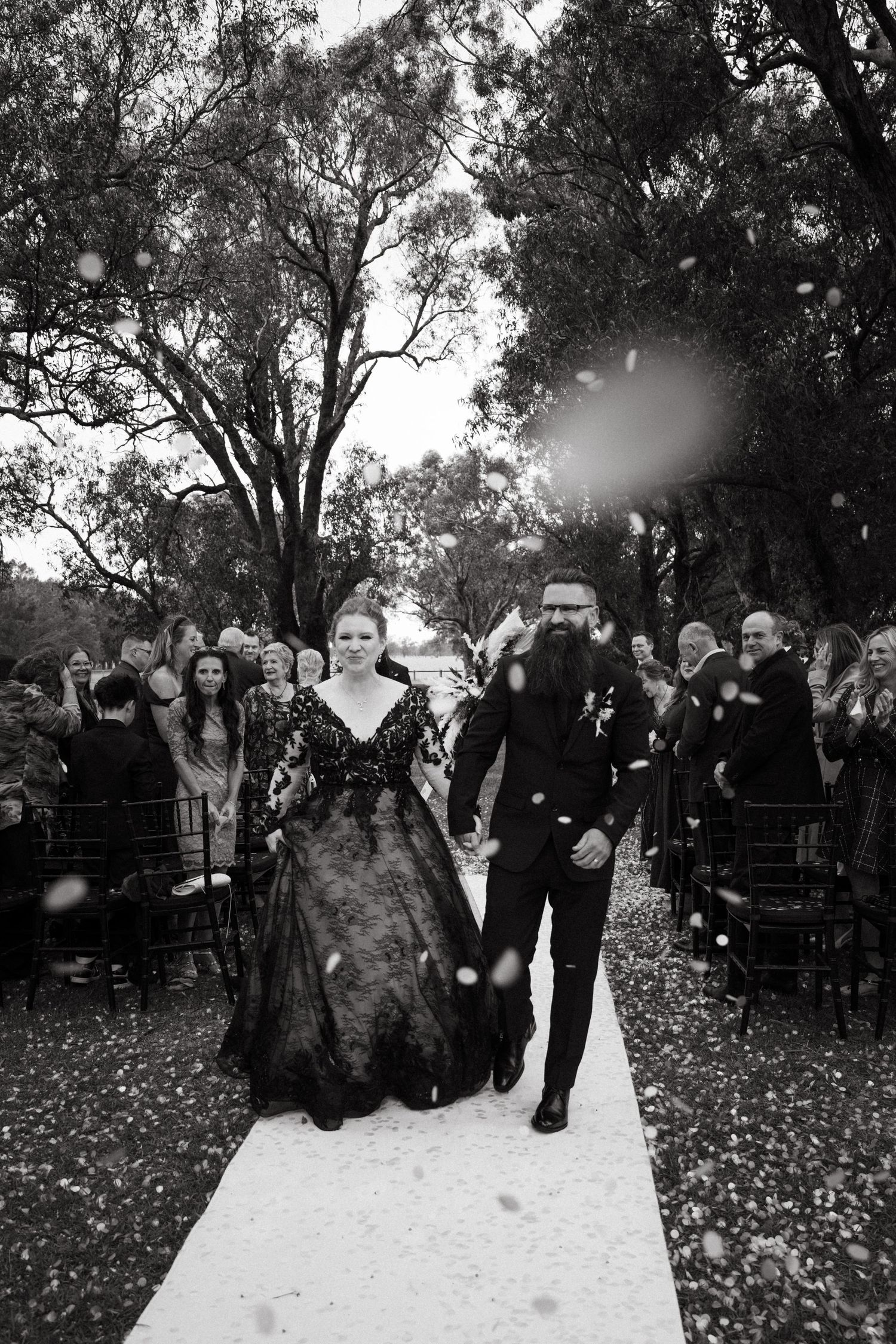Black and white photo of newlyweds walking down the aisle in black wedding attire, surrounded by guests throwing confetti.