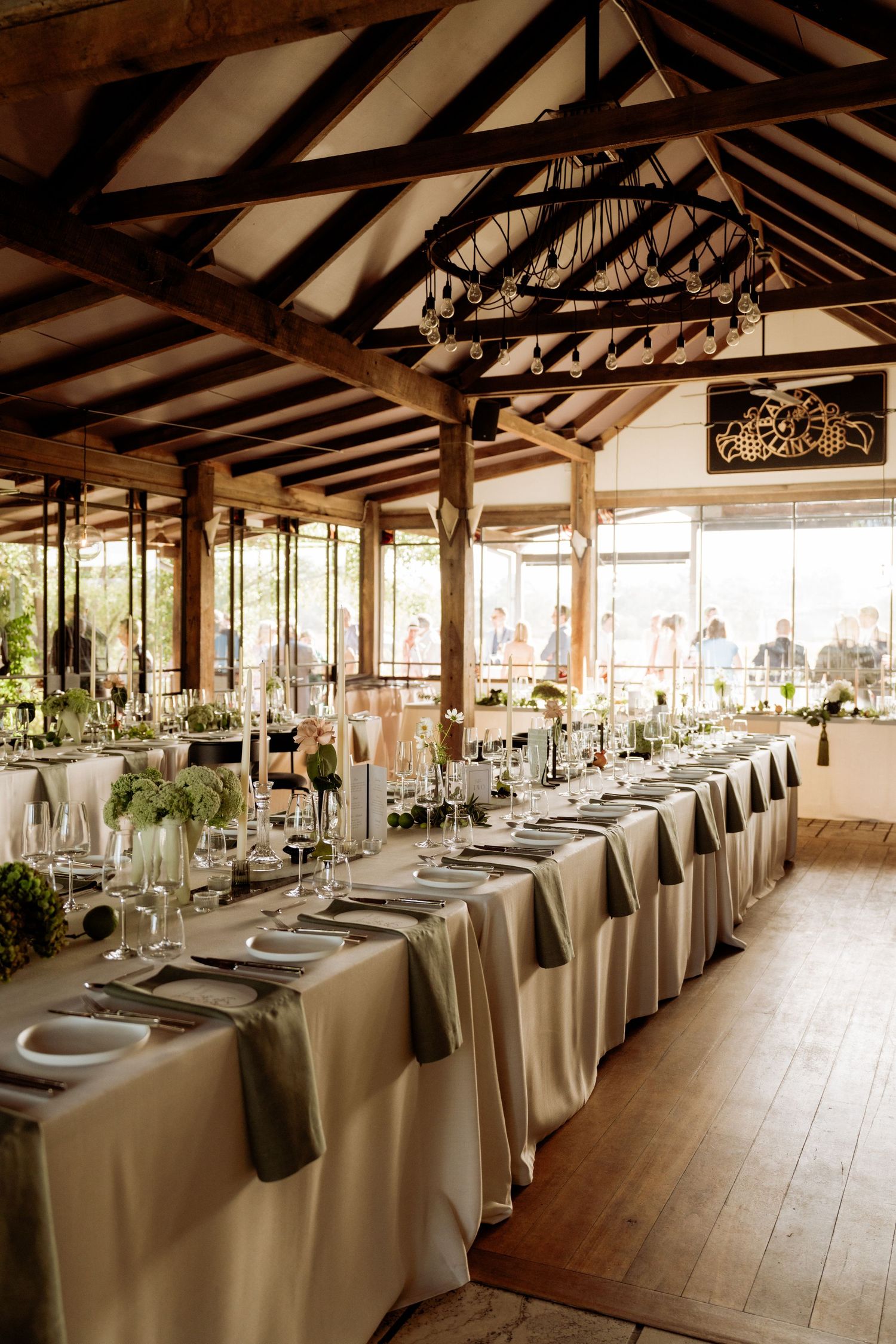 Elegant wedding reception hall with long tables, white linens, gray napkins, floral centerpieces, and rustic wooden beam ceiling.