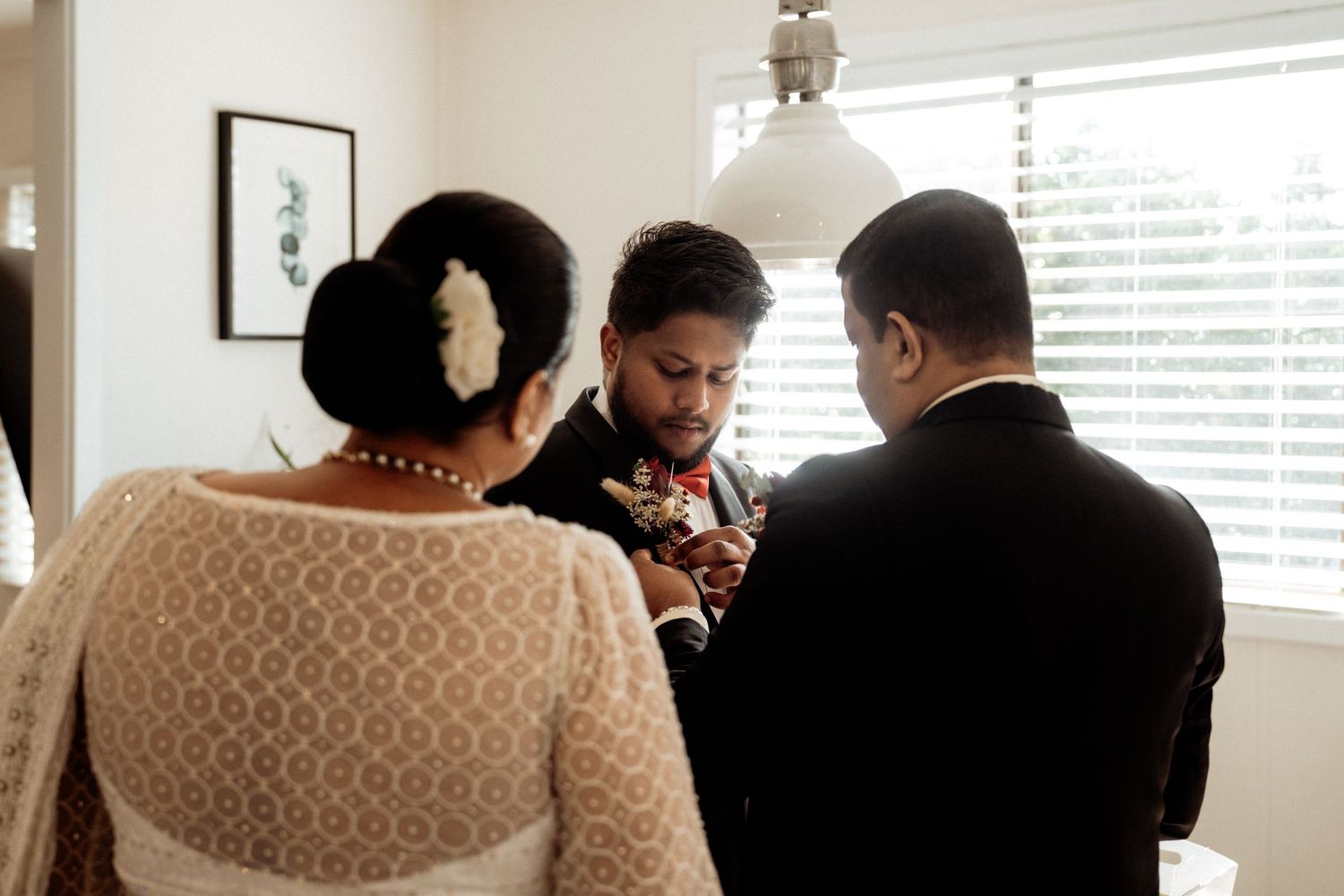 Three people in formal attire gather indoors near window blinds during a wedding preparation ceremony.