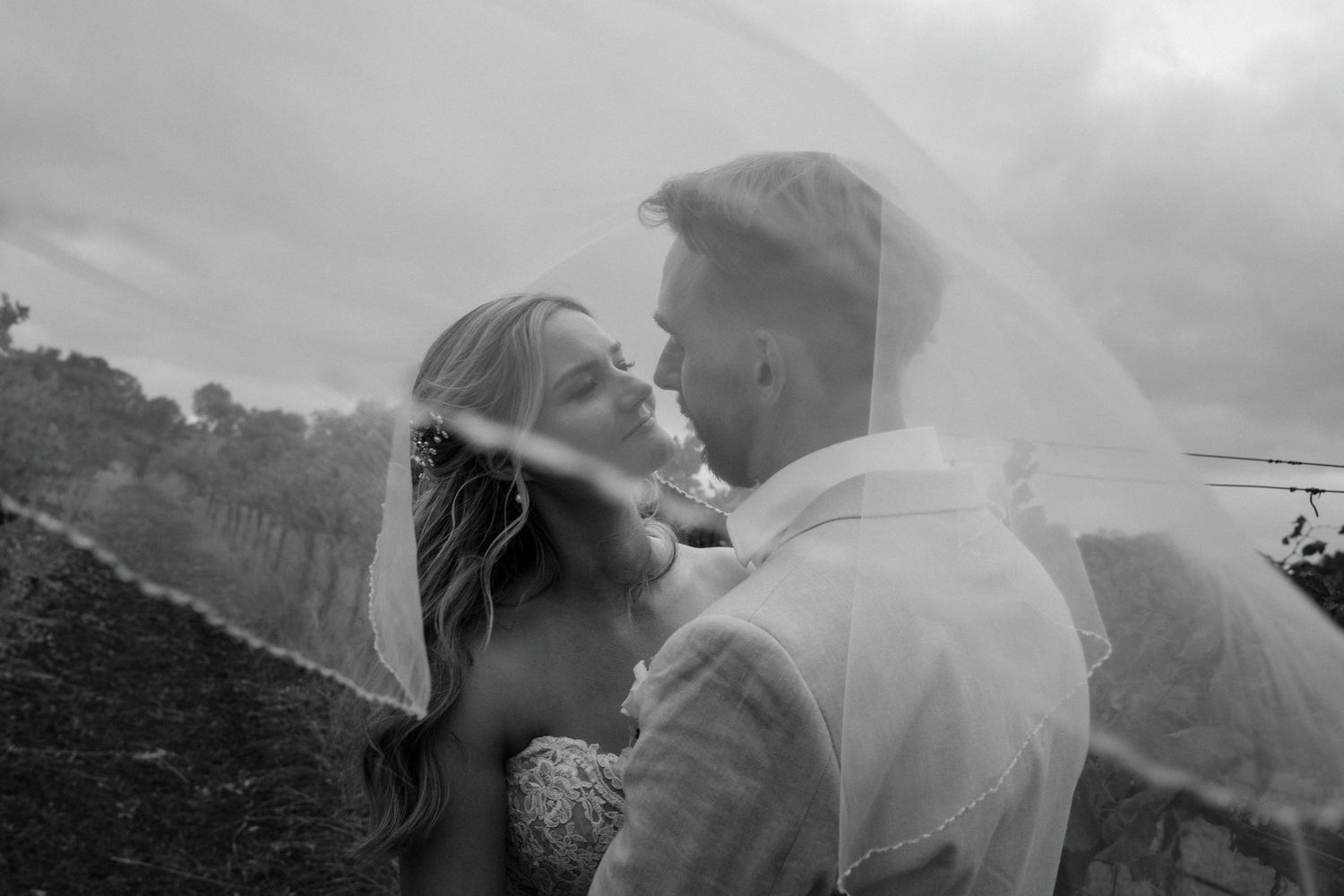 Black and white wedding photo of bride and groom sharing intimate moment under flowing veil outdoors.