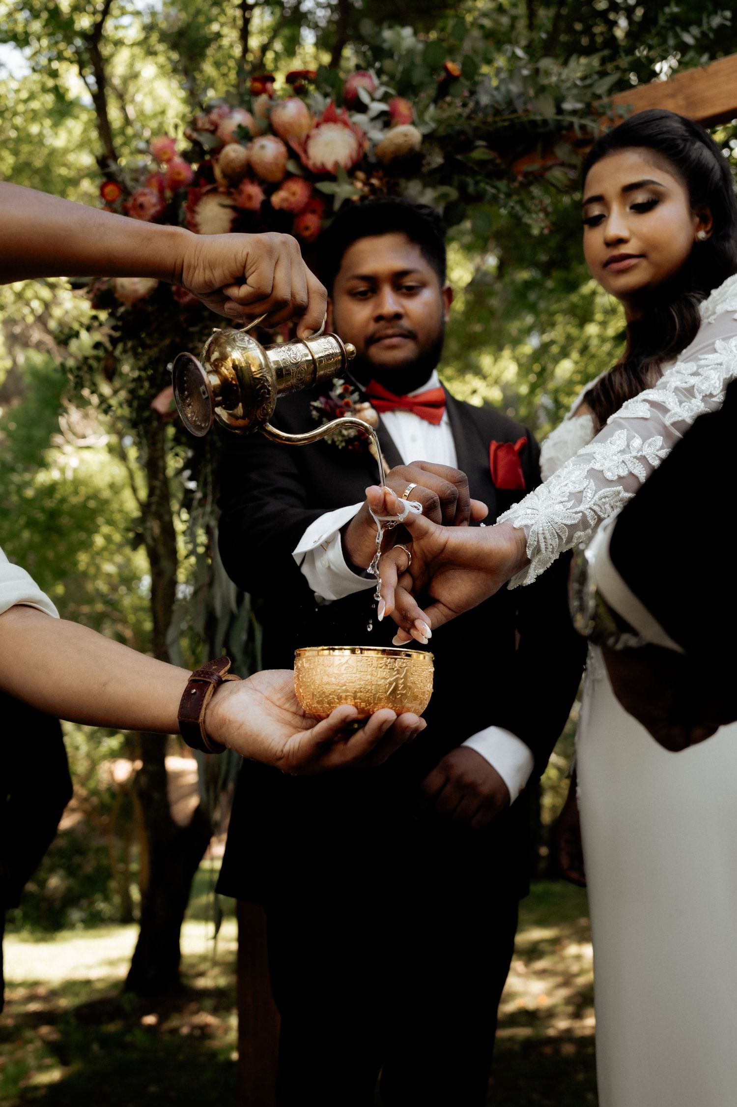 Bride and groom perform a traditional water ritual ceremony outdoors, with a golden bowl and ornate brass pitcher.