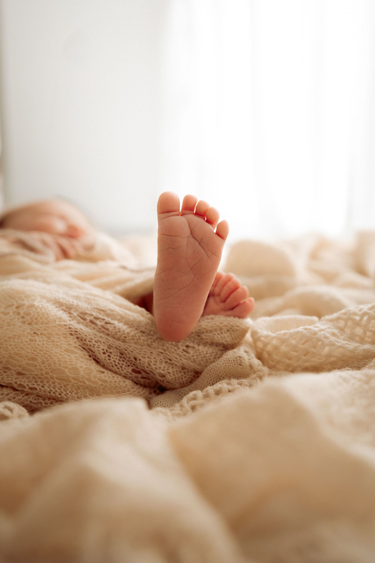 Close-up of tiny baby foot resting on soft beige blankets in natural window light.