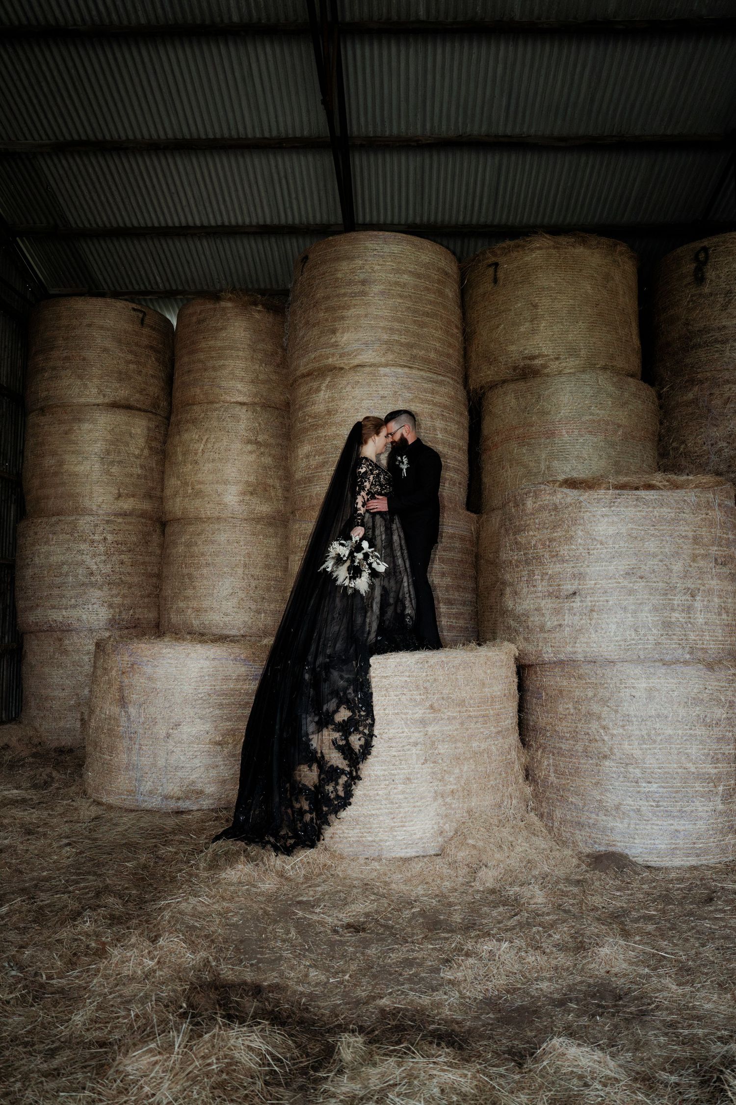 Couple in black wedding attire kissing among stacked hay bales in a rustic barn setting.