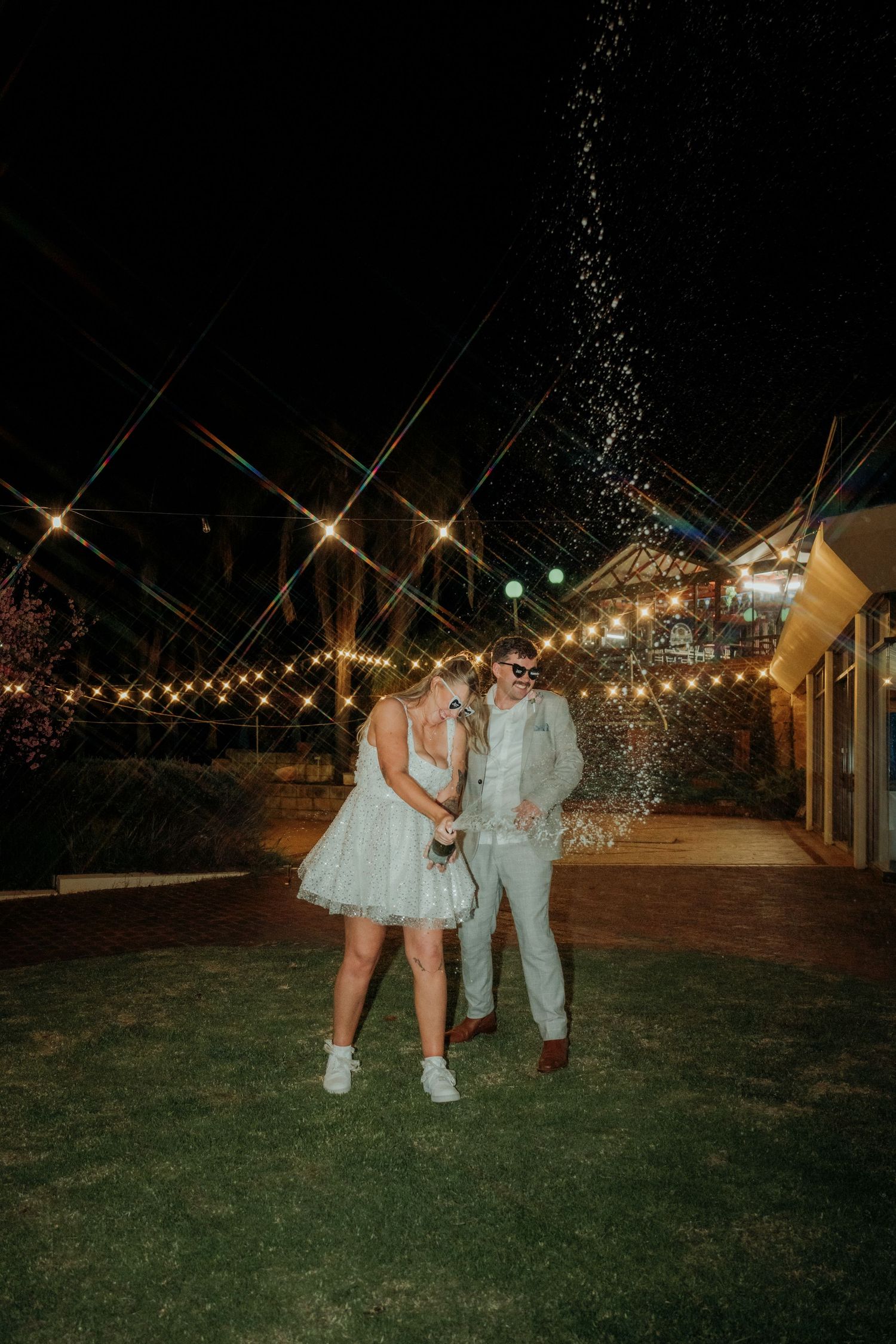 Couple in white attire celebrating outdoors at night under string lights with champagne spray.