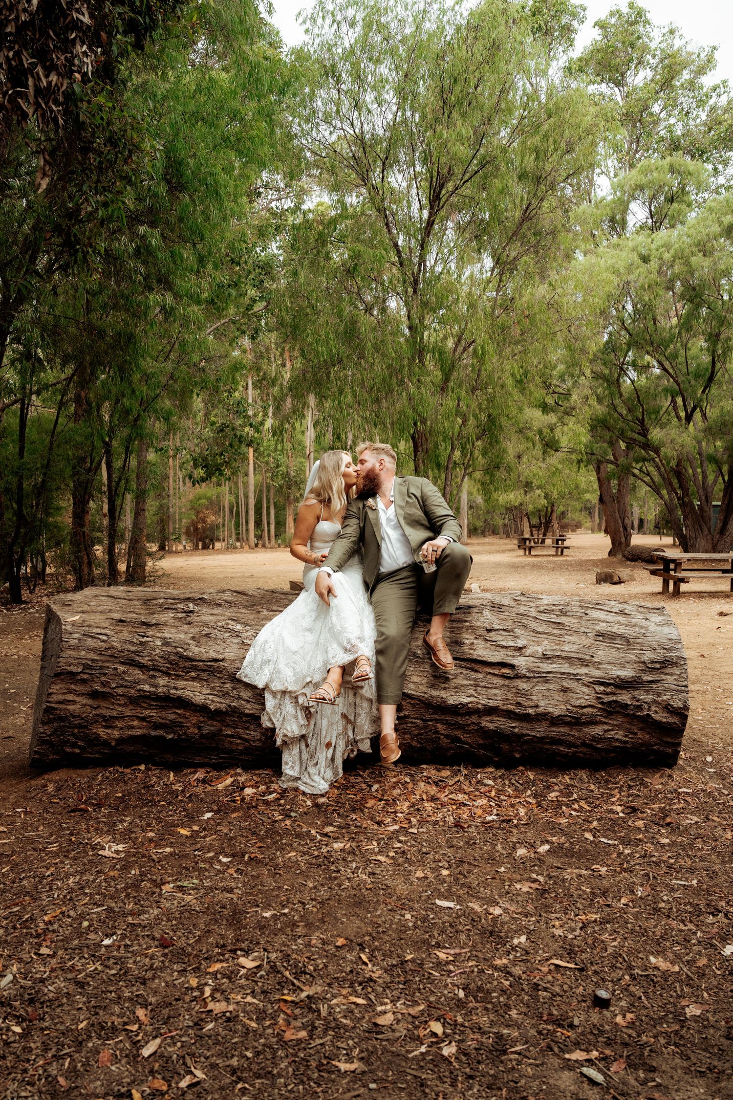Bride and groom sharing a romantic kiss while sitting on a fallen log in a lush forest setting.