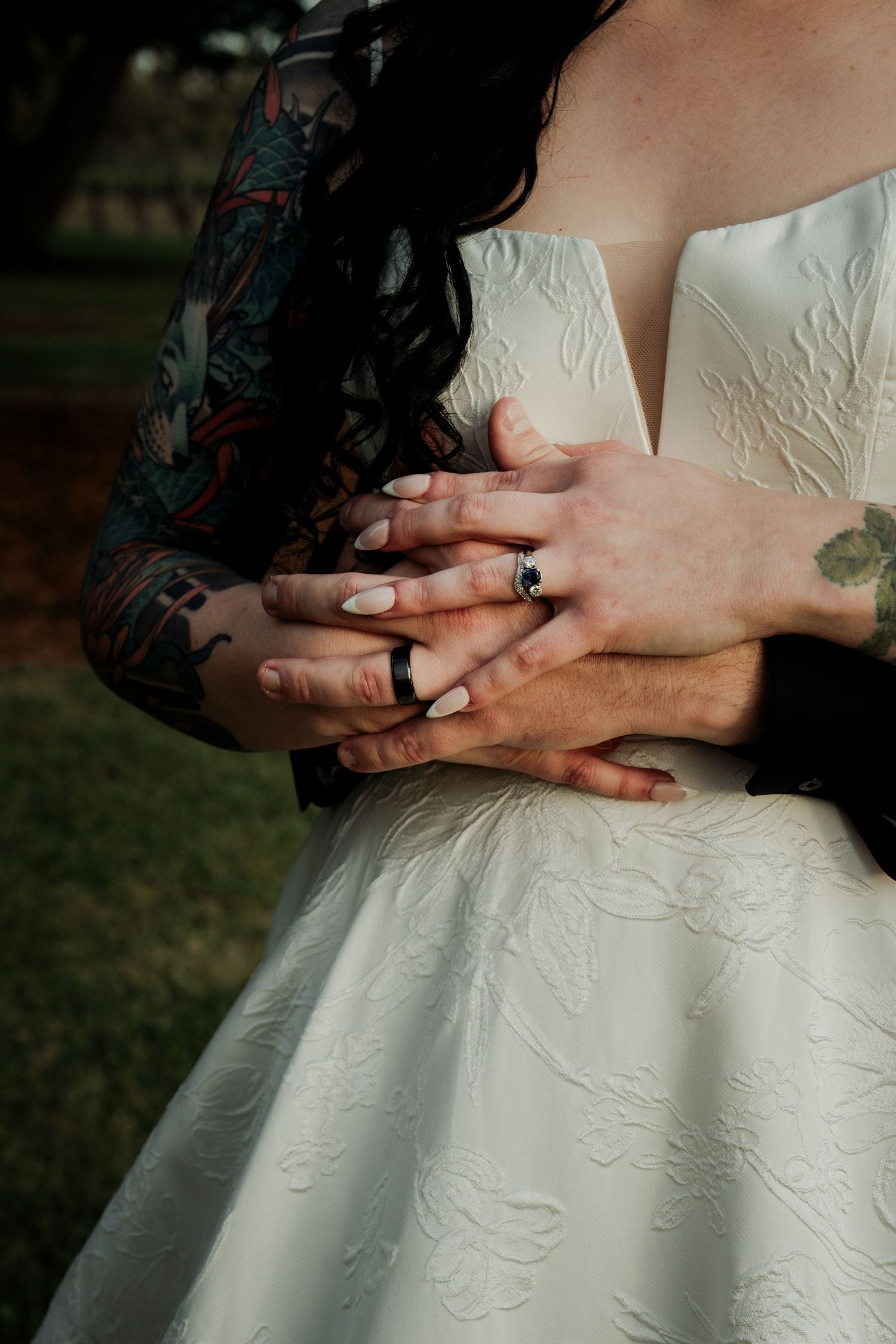 Bride and groom showing wedding rings, groom with colorful tattoo sleeve embracing bride in white gown.