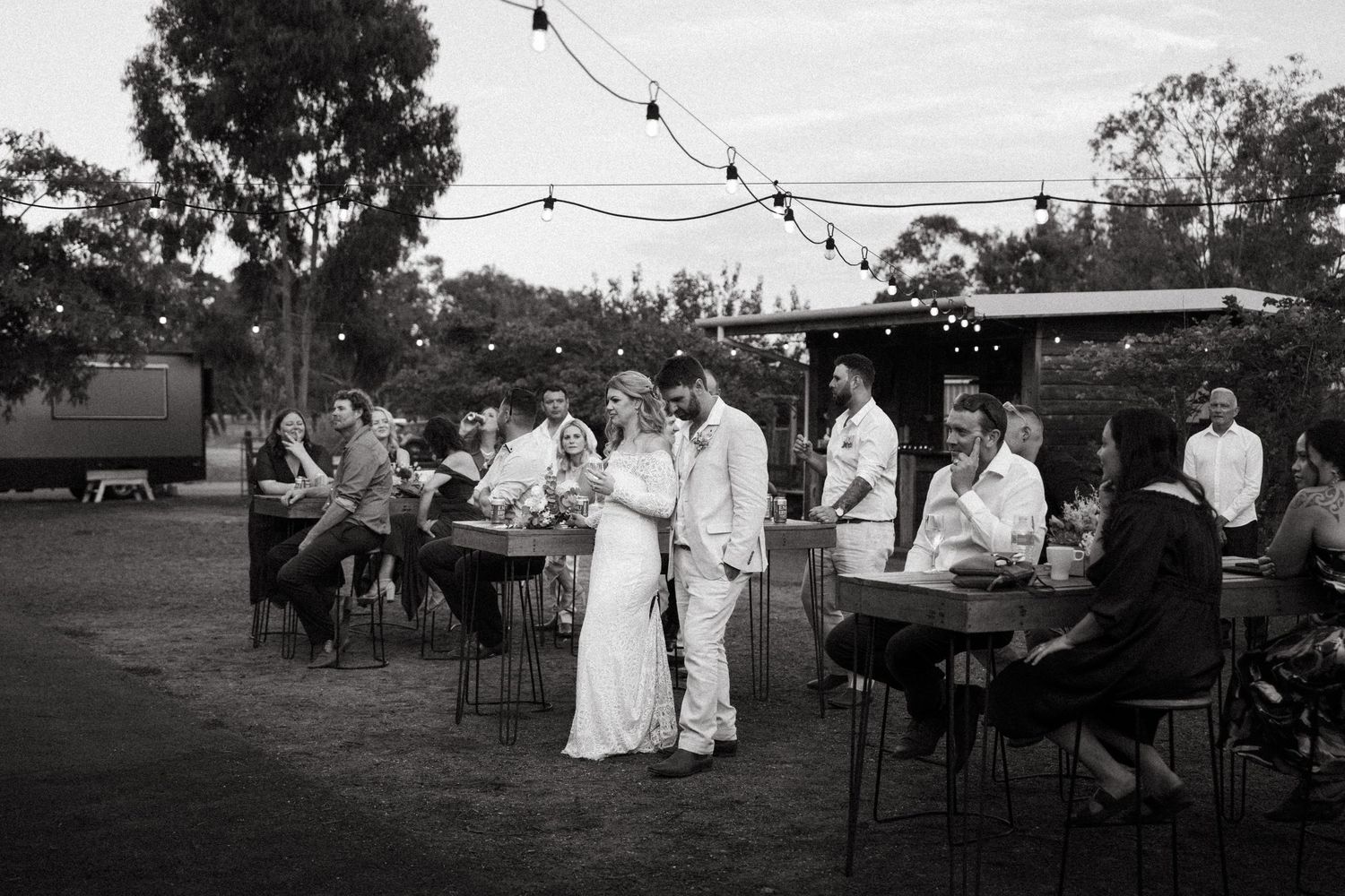 Black and white photo of wedding guests mingling outdoors under string lights at a rustic reception.