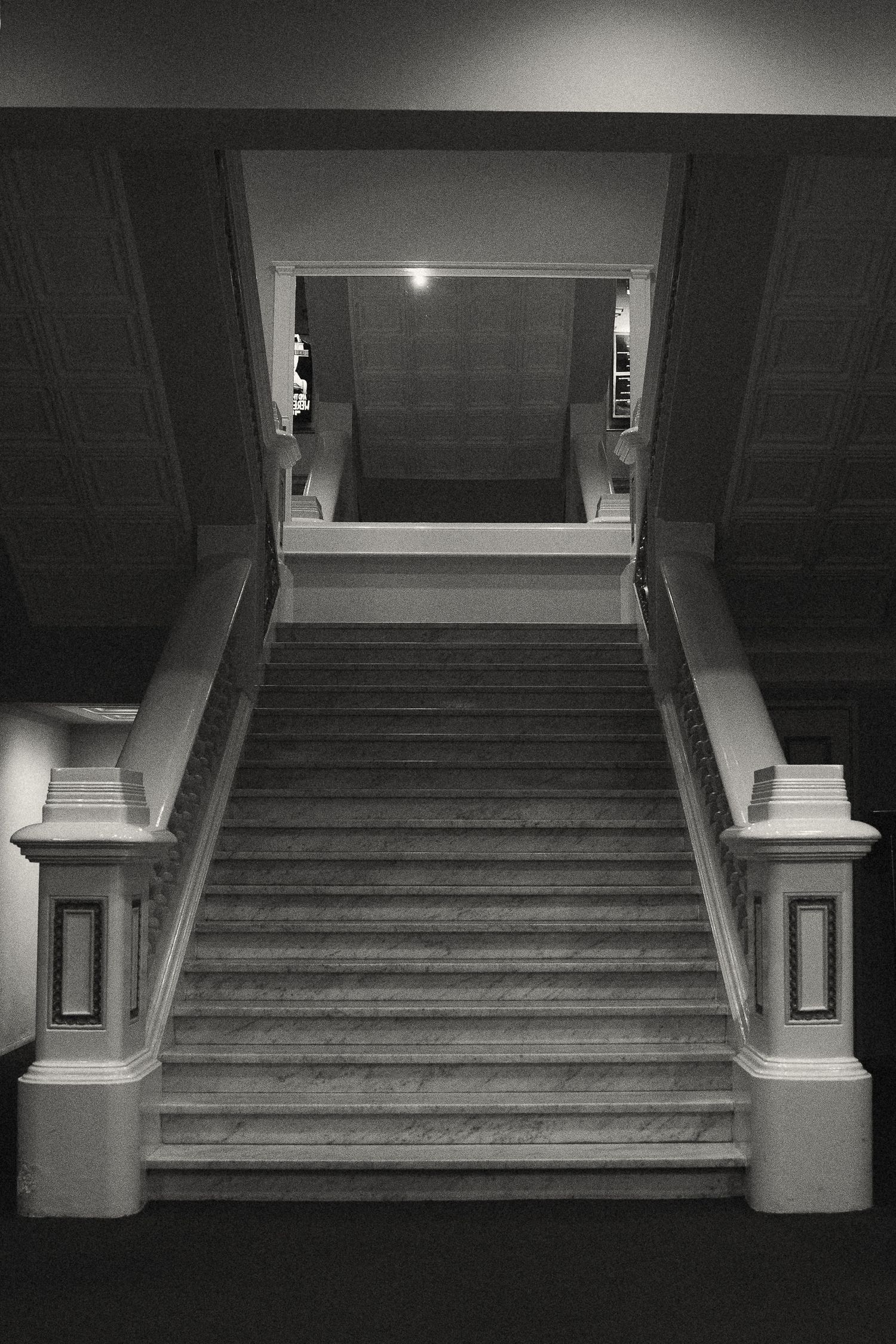 A dramatic black and white photograph of a grand staircase with ornate pillars and railings in low-key lighting.