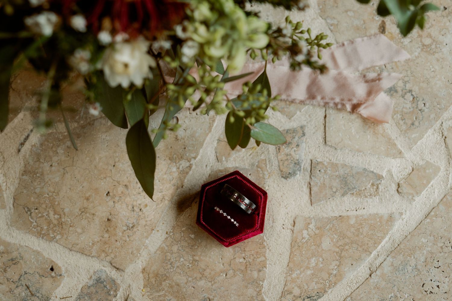 Wedding rings displayed in a red velvet box on stone surface with bridal bouquet of greenery and white flowers in background.