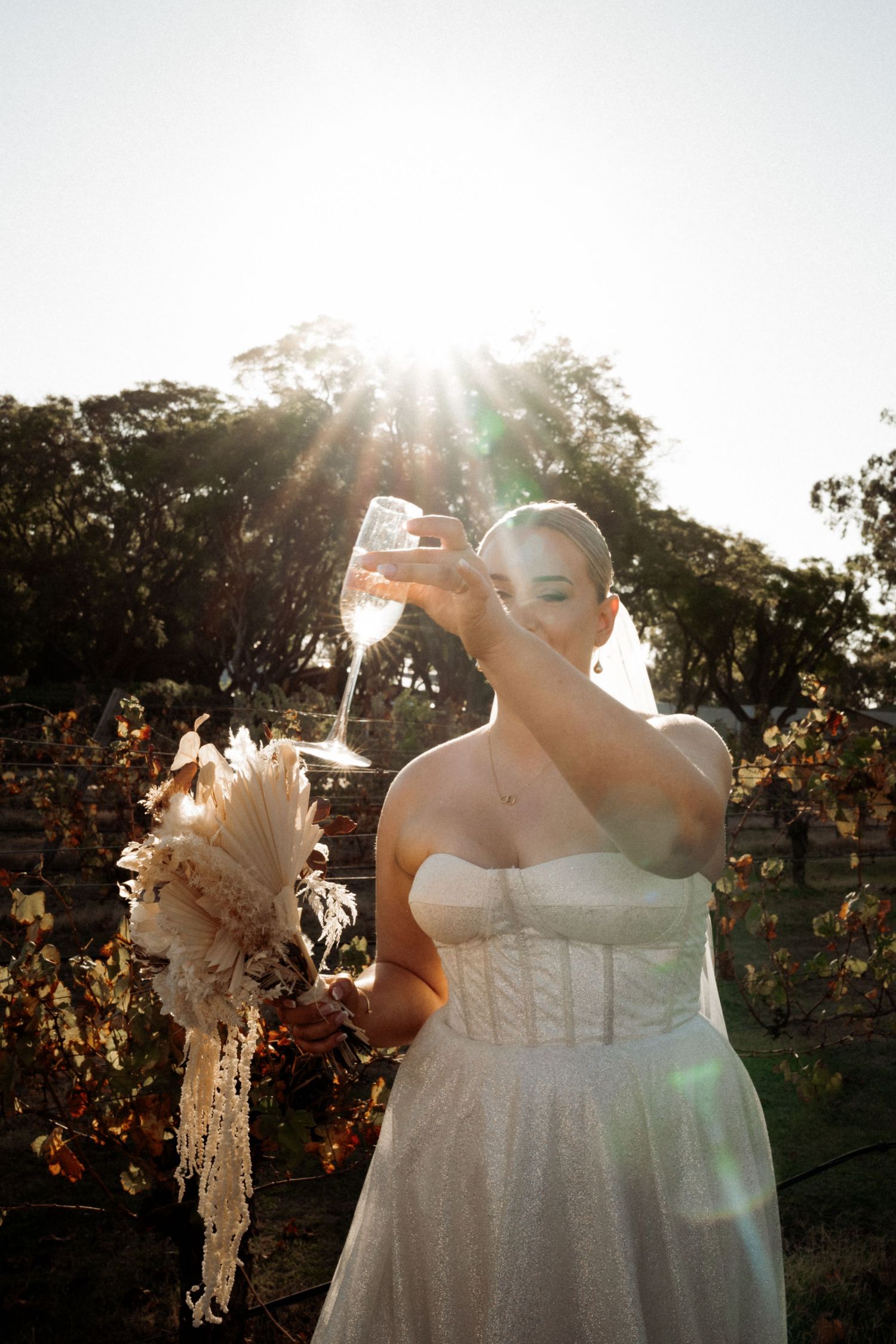 Bride in strapless white gown drinking wine at sunset vineyard, holding dried floral bouquet with golden lens flare.