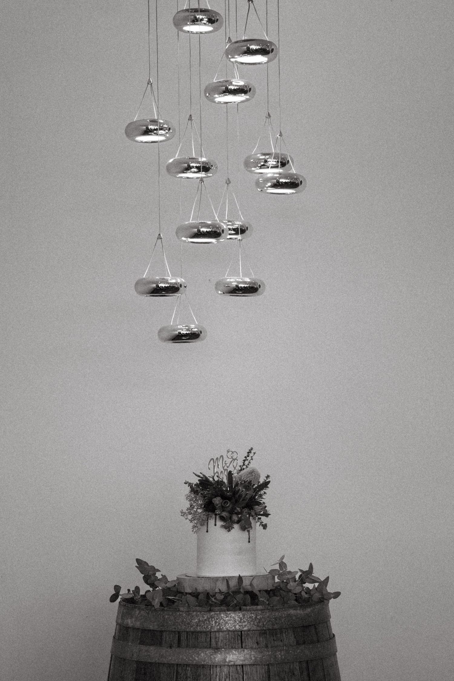 Black and white photo of a wedding cake on a wine barrel beneath cascading metallic pendant lights.