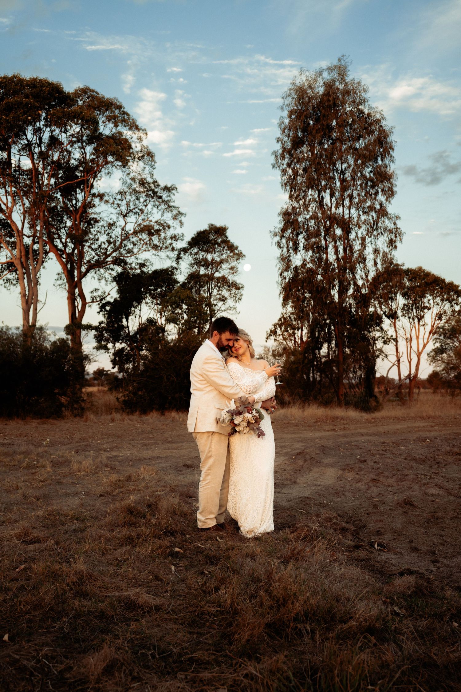 Bride and groom embrace in golden hour light in a rustic field with eucalyptus trees, bride holding a bouquet.