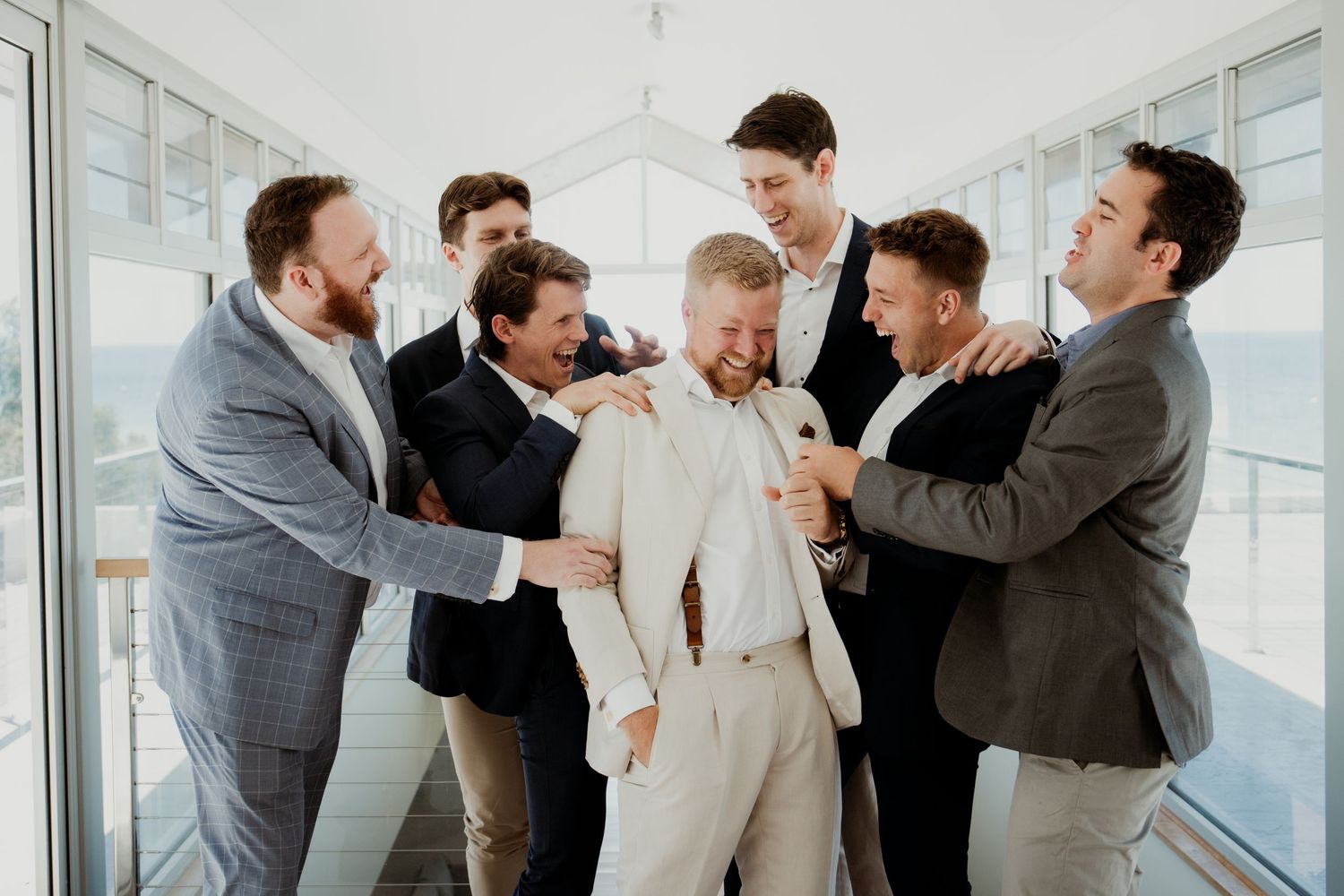 Groom in white suit laughing as groomsmen in gray suits celebrate together in bright indoor venue.