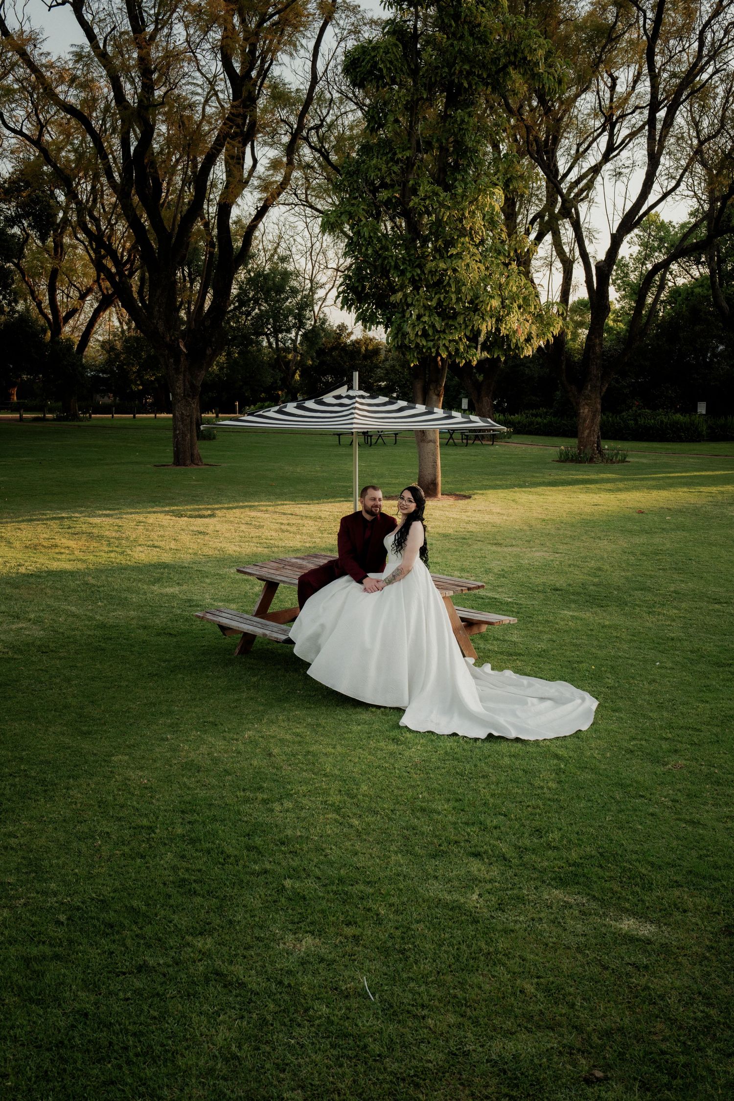 Bride and groom sitting together at a picnic table under a striped umbrella in a scenic park wedding setting.