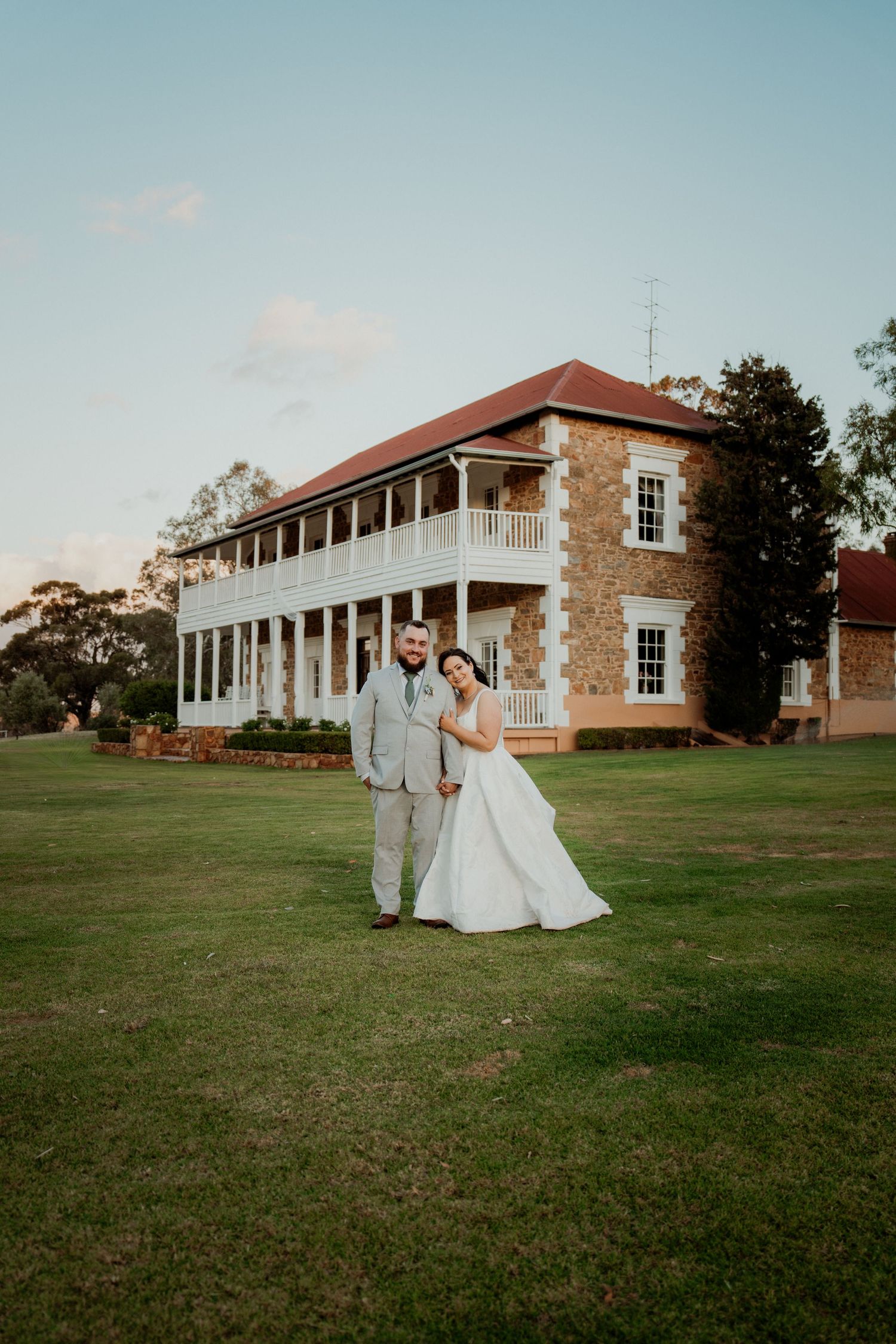 Bride and groom pose on green lawn before historic two-story stone manor with red roof on their wedding day.