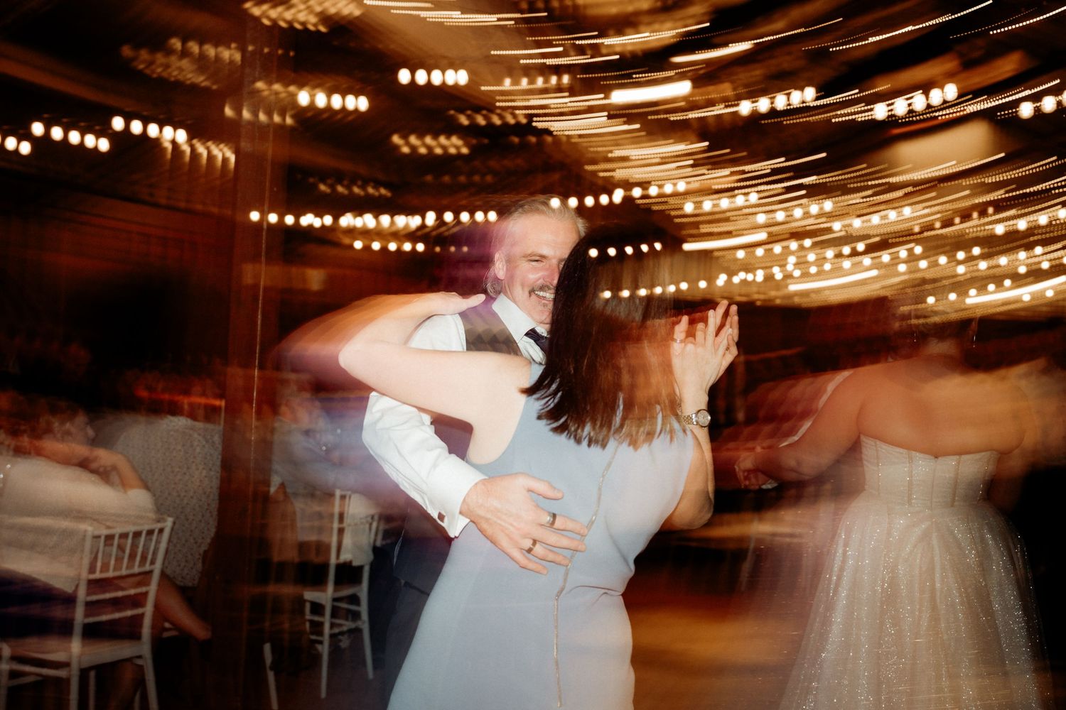 Couple shares romantic first dance at wedding reception with warm string lights swirling in background.