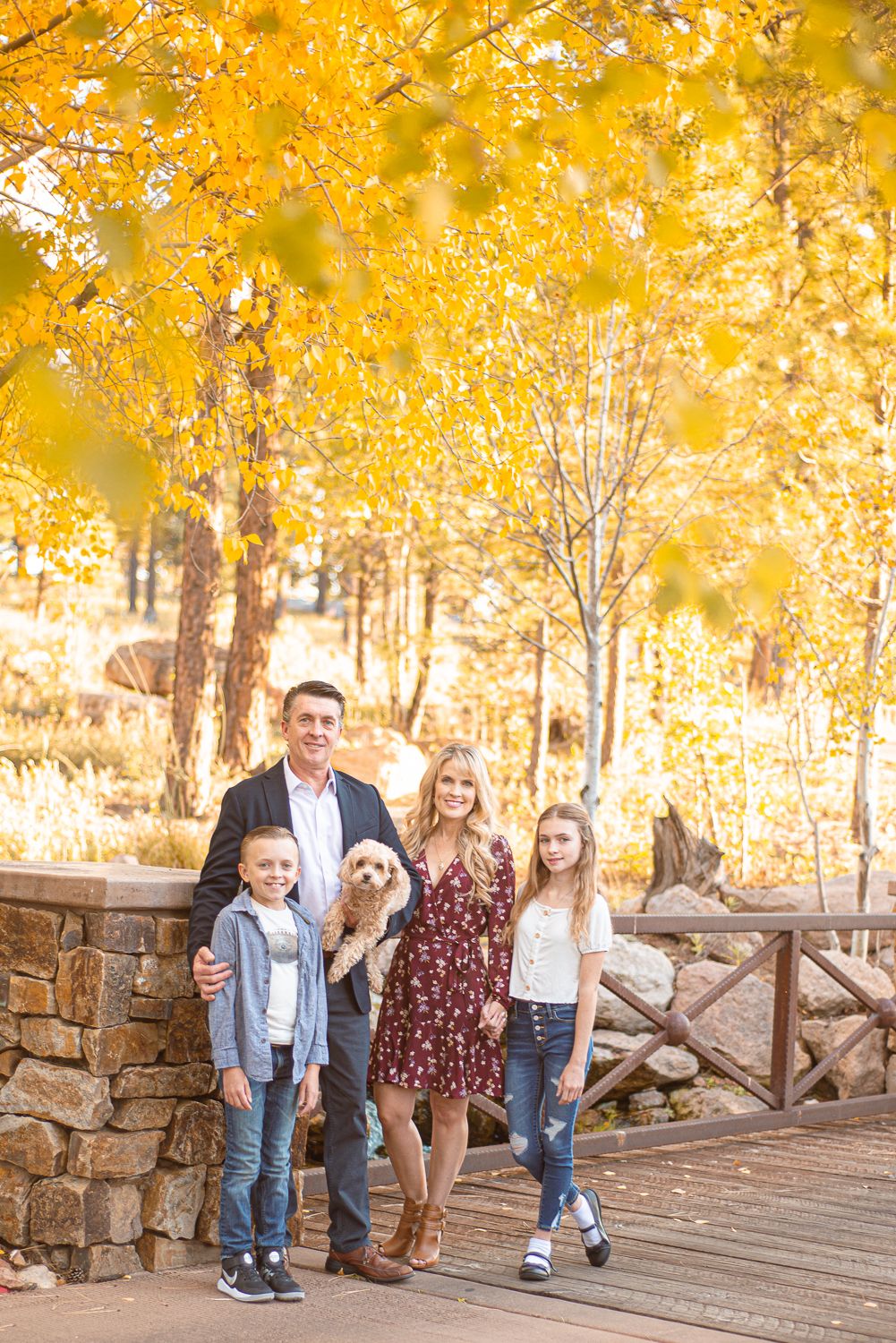 Family of four with a dog posing on a bridge surrounded by golden autumn foliage trees.