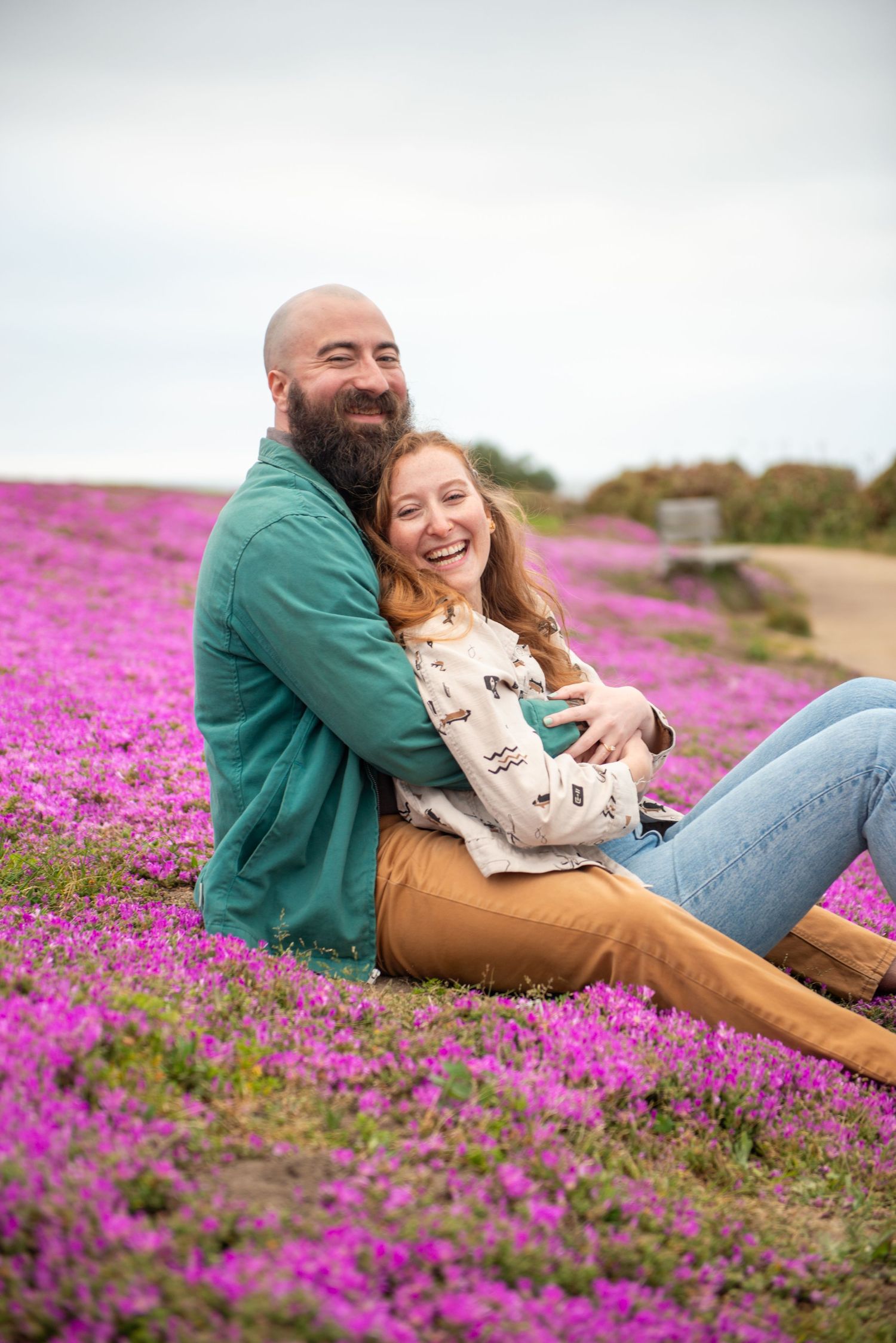 Couple laughing and embracing while sitting in a vibrant purple flower field outdoors.