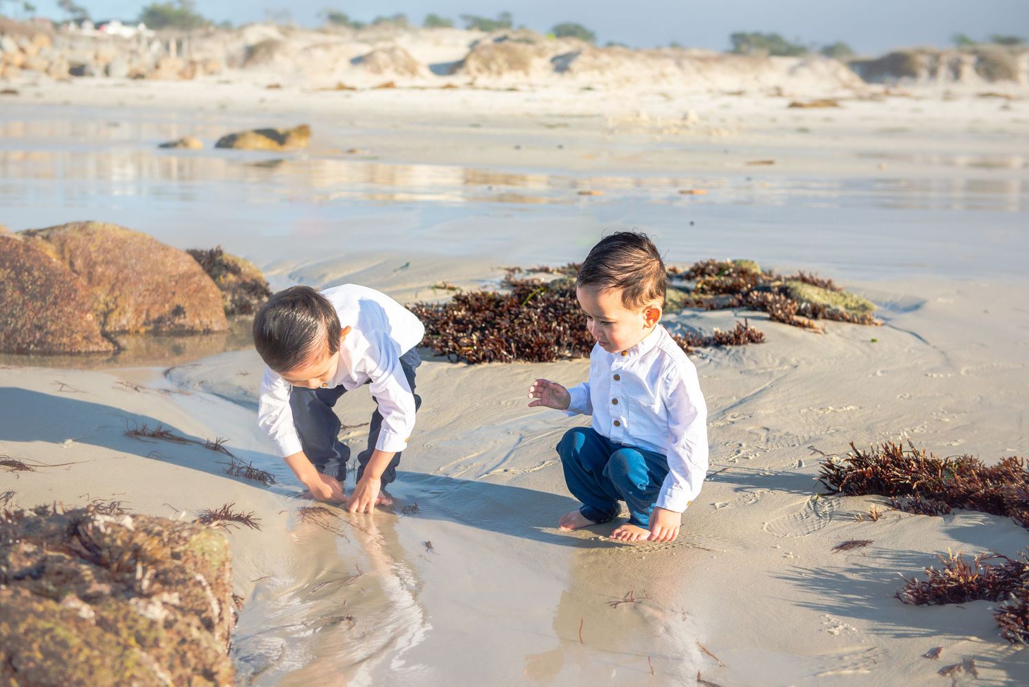 Two children crouching to play in the wet sand at the beach with seaweed and rocks nearby during a sunny day.