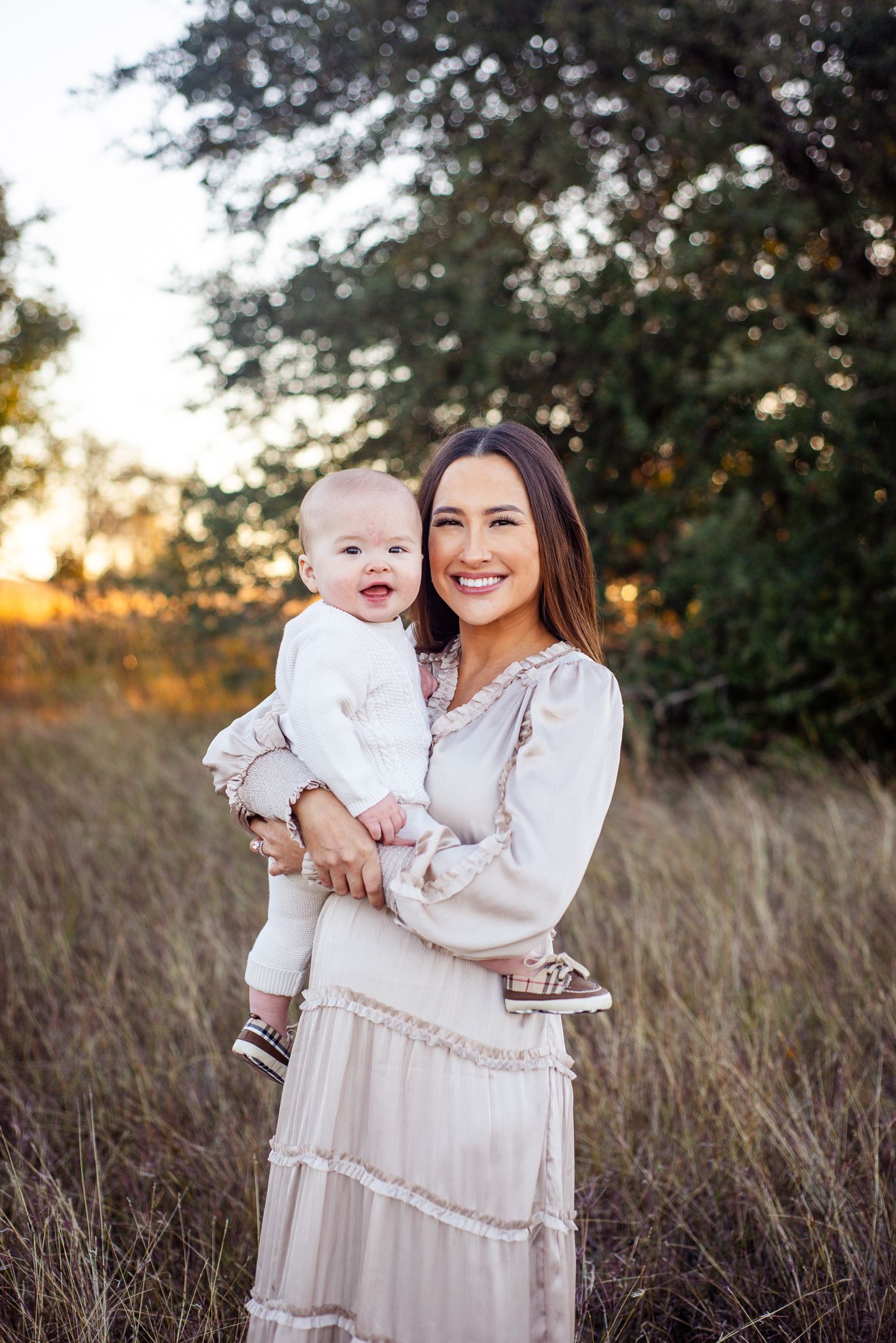 Beautiful outdoor portrait of parent and child in matching white dresses standing in tall grass at golden sunset.