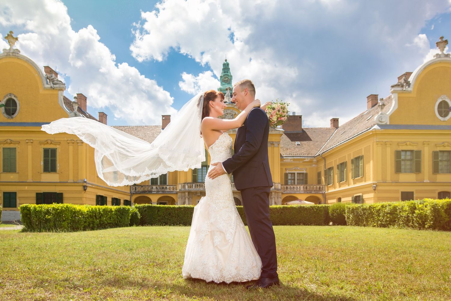 A couple embraces on the lawn in front of elegant yellow baroque buildings as the bride's veil flows dramatically in the wind.