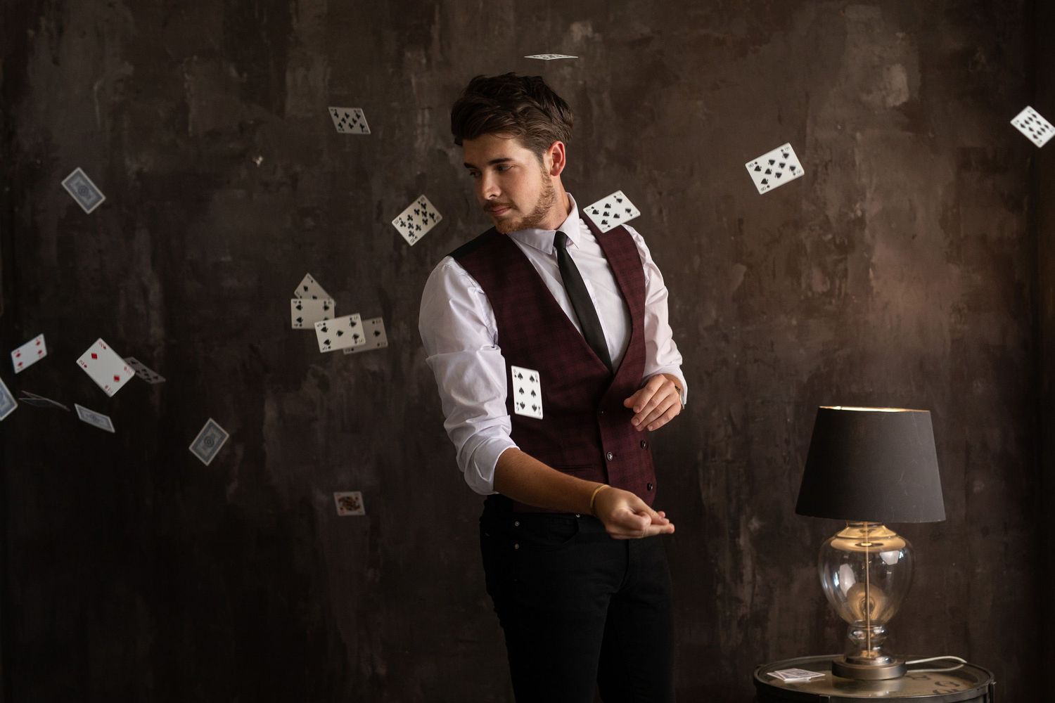 A magician performs card tricks against a dark wall with playing cards floating in the air next to a table lamp.