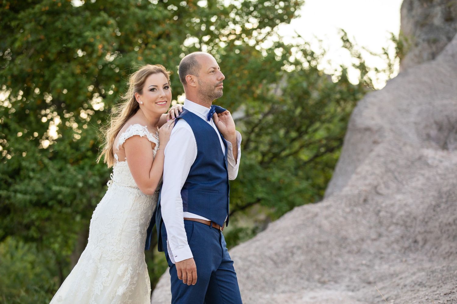 Bride and groom walk together on outdoor path at sunset surrounded by green foliage and stone formations.
