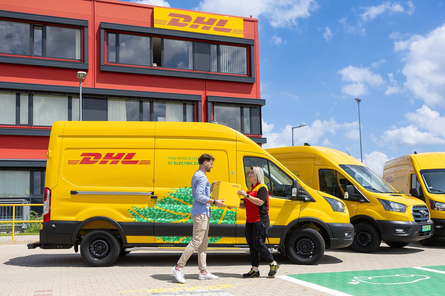 Fleet of yellow DHL delivery vans parked on green pavement with buildings visible in the background.