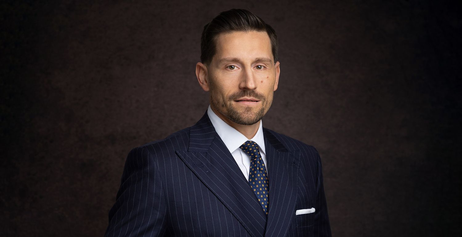 Professional headshot portrait of a person in a dark navy suit and tie against a dramatic black background.