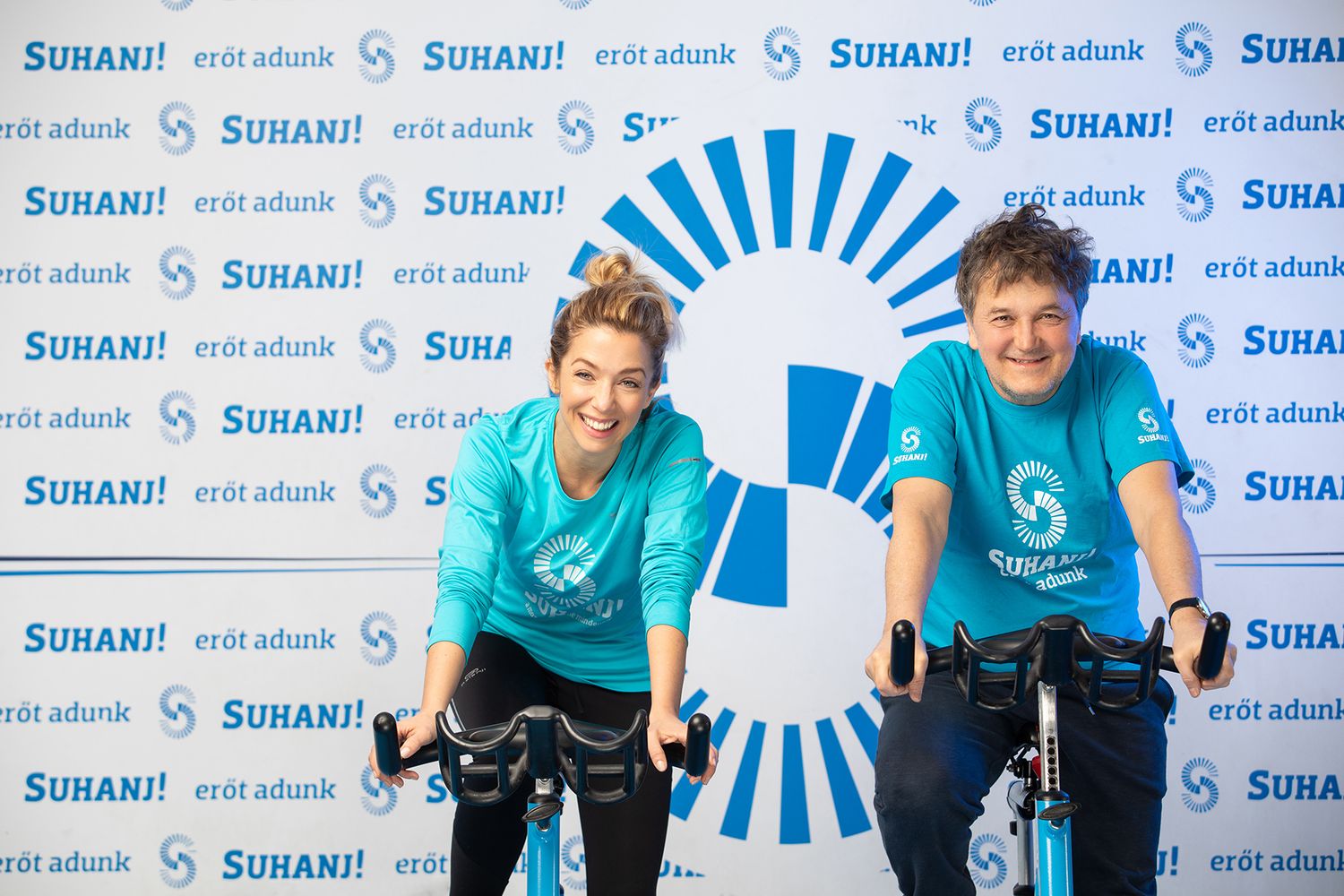 Two people pose with Sunami branded exercise bikes at a promotional event with repeating blue logos on the backdrop.